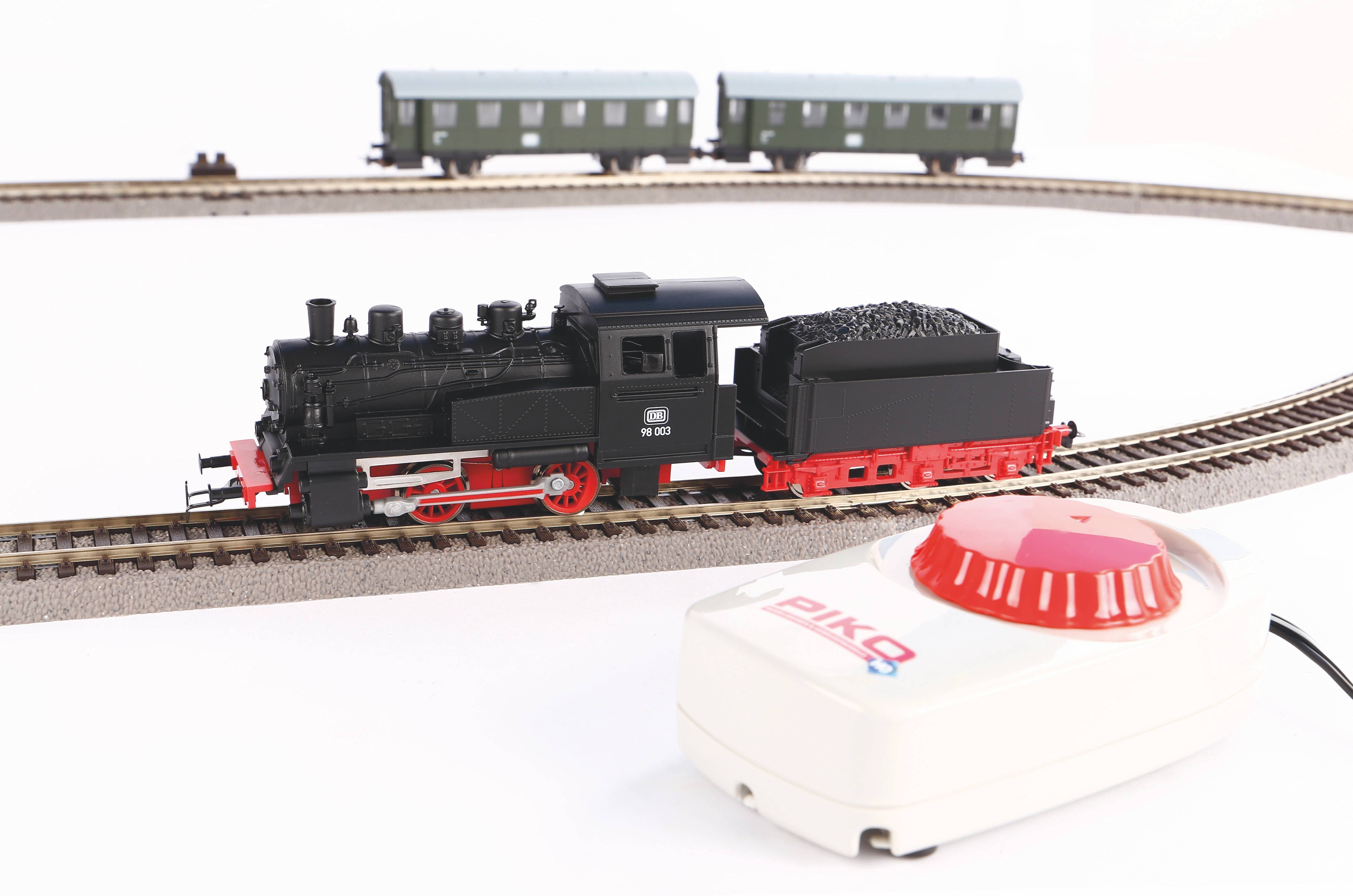 Een modeltreinstel op een rail, met een zwarte locomotief en twee groene wagons op de achtergrond, naast een besturingsapparaat.