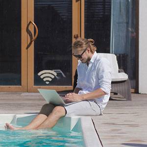 Een man zit met een laptop bij het zwembad. Hij draagt een wit overhemd en een zonnebril. Op de achtergrond is een glazen deur zichtbaar.