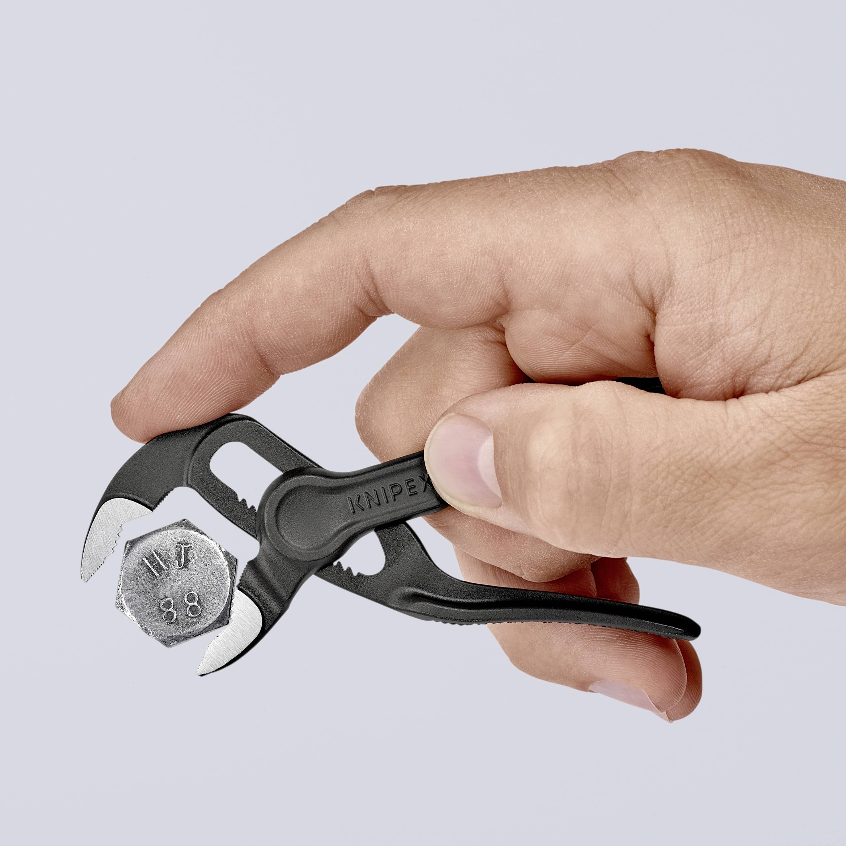 Een hand houdt een kleine, zwarte tang vast die een zeskantmoer vastgrijpt. De tang is compact en handzaam gebouwd.