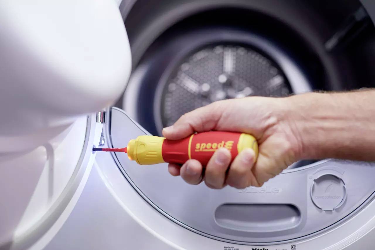 Een hand houdt een gele en rode schroevendraaier vast en draait een schroef vast aan de deur van een wasmachine.