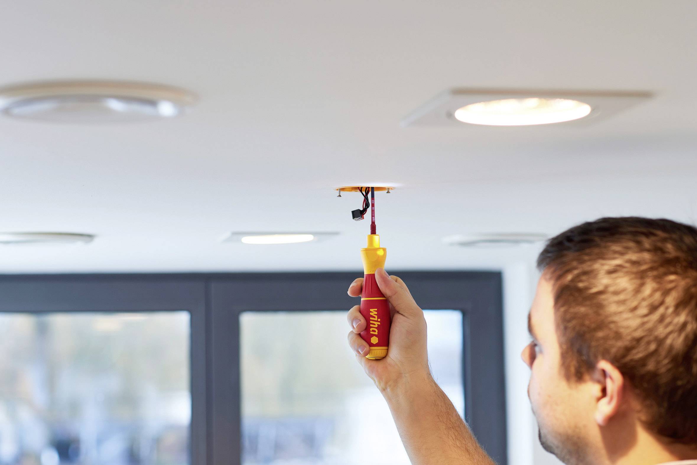 'Persoon installeert plafondverlichting met schroevendraaier met gele handgreep. Op de achtergrond zijn ramen zichtbaar.'
