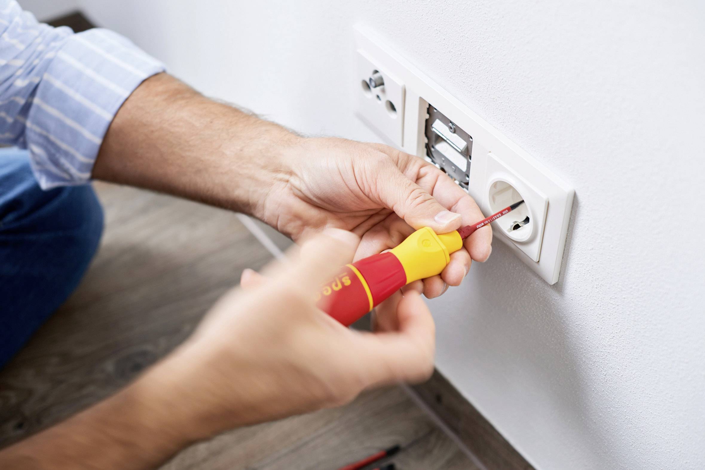 Een persoon repareert een wandcontactdoos met een schroevendraaier. De schroeven worden afgesteld. Het gaat om elektrische werkzaamheden.