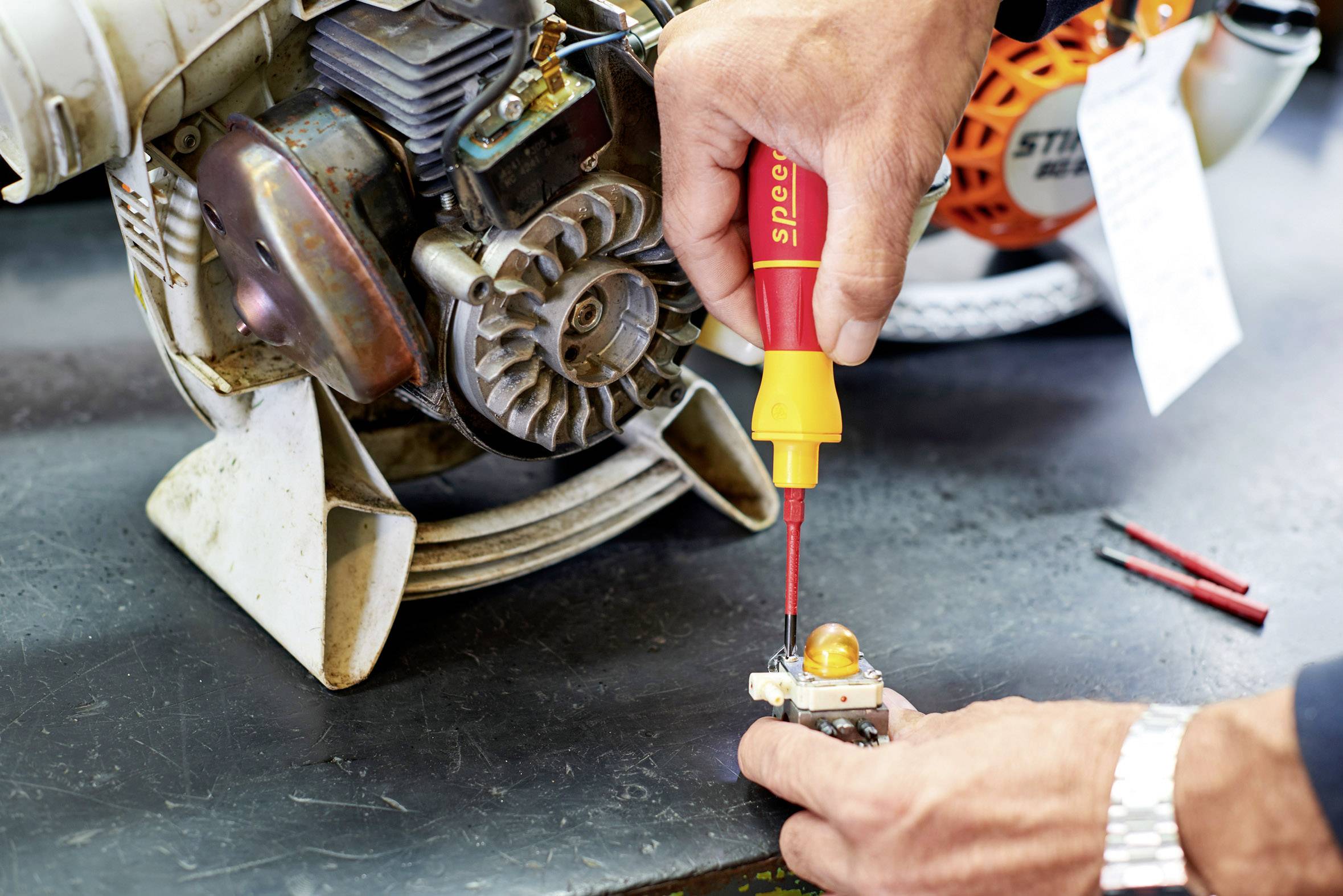 'Een persoon repareert een motor met een schroevendraaier, close-up van de motor en de handen. Op tafel liggen nog meer gereedschappen.'