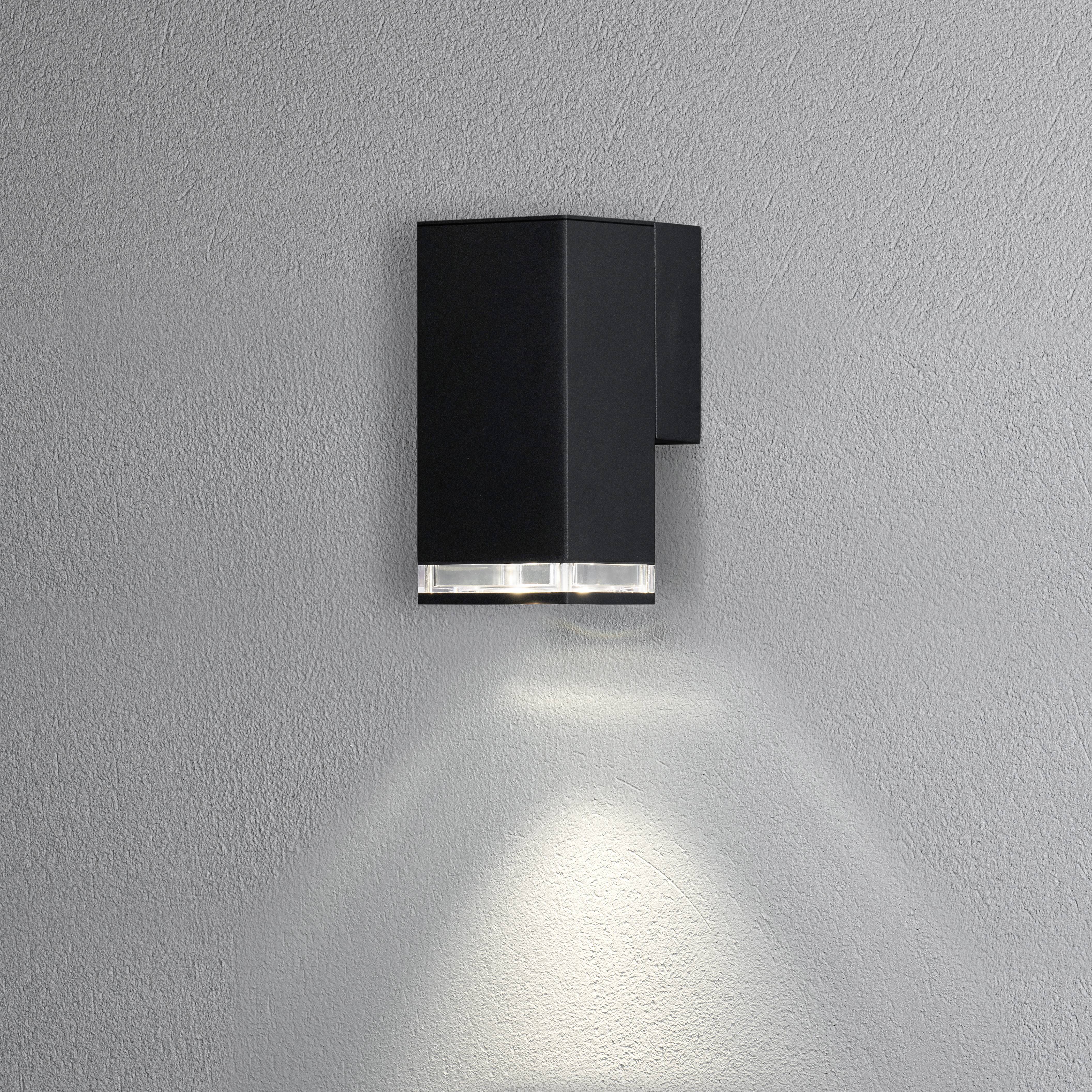 Wandlamp in modern, kubistisch design met zwarte behuizing, schijnt naar beneden en creëert zacht licht op grijze muur.