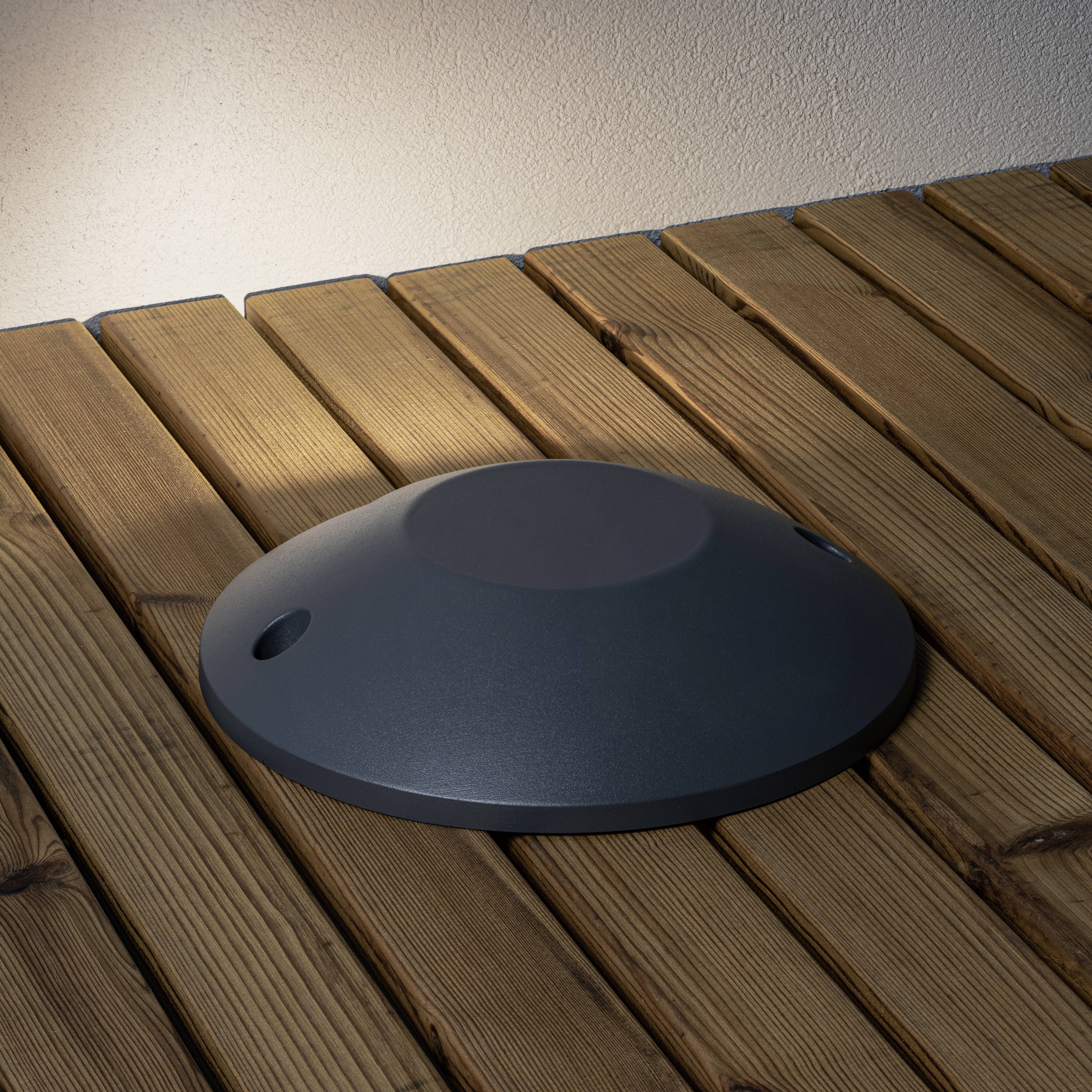 Een donkere, platte lamp schijnt op een houten terras, verlicht een muur op de achtergrond.