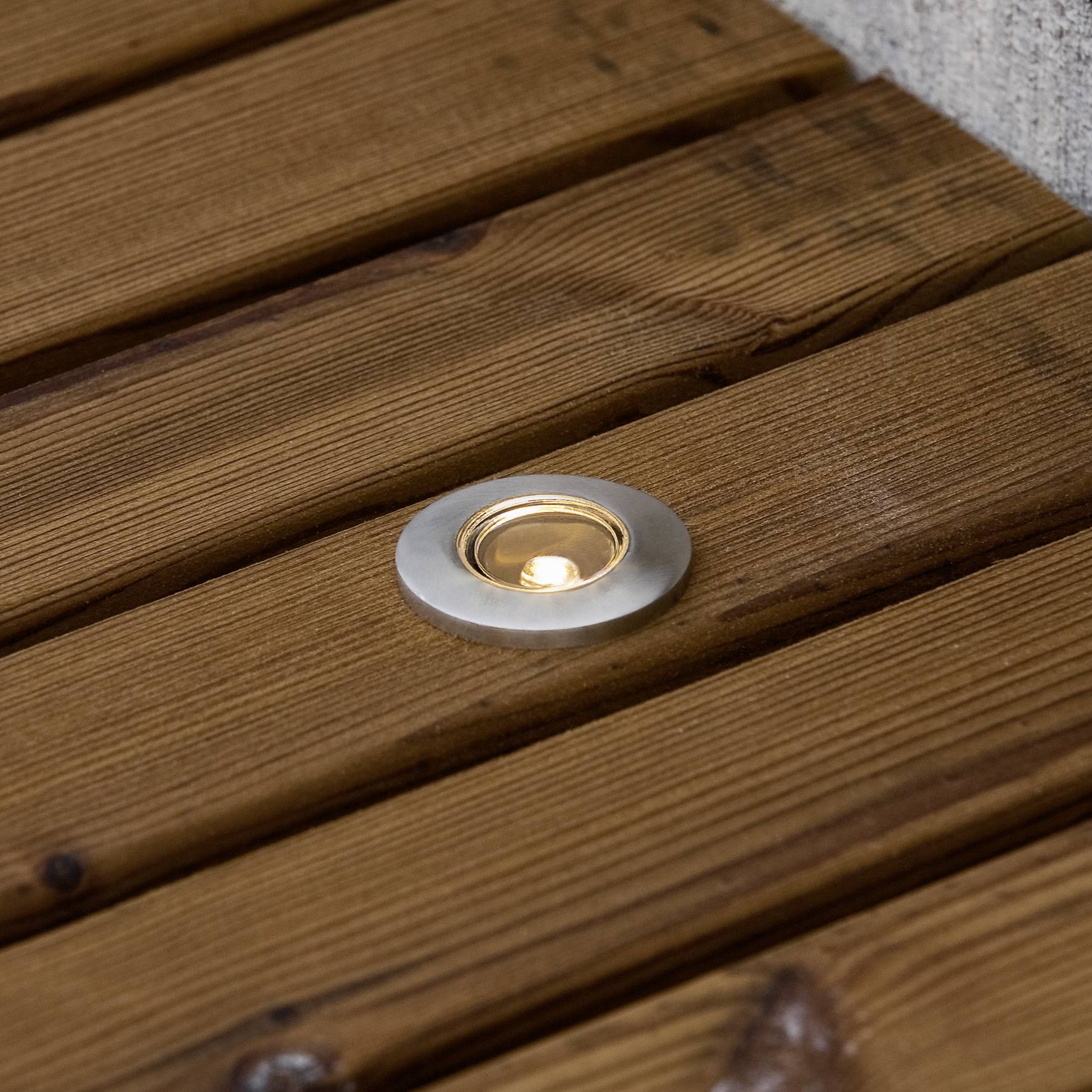 Een ronde inbouwspot is verzonken in een houten terrasvloer en straalt een warme gloed uit.