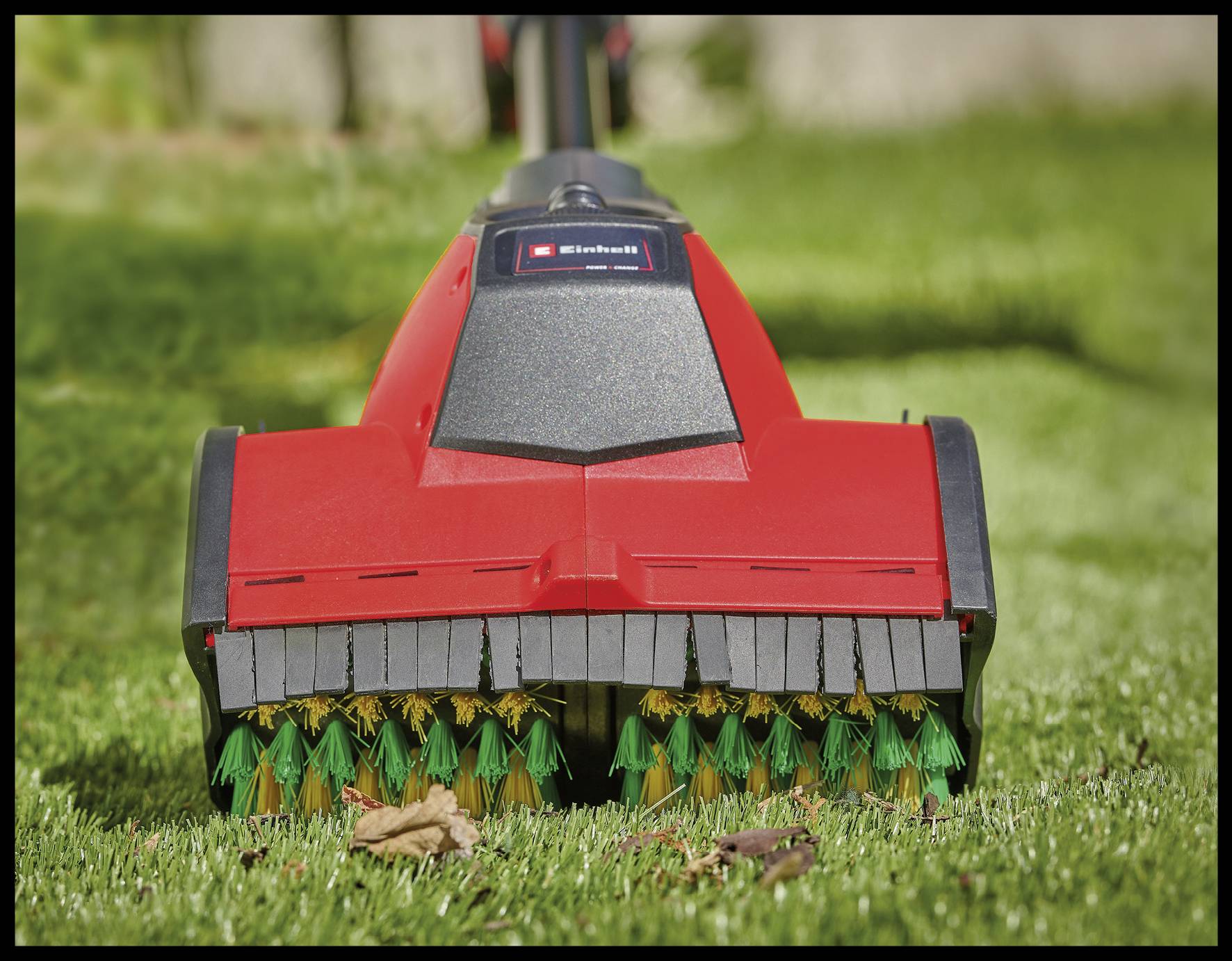 Een rode verticuteermachine werkt op een groene gazonomgeving, waarbij roterende borstels zichtbaar zijn die mos en dood gras verwijderen.