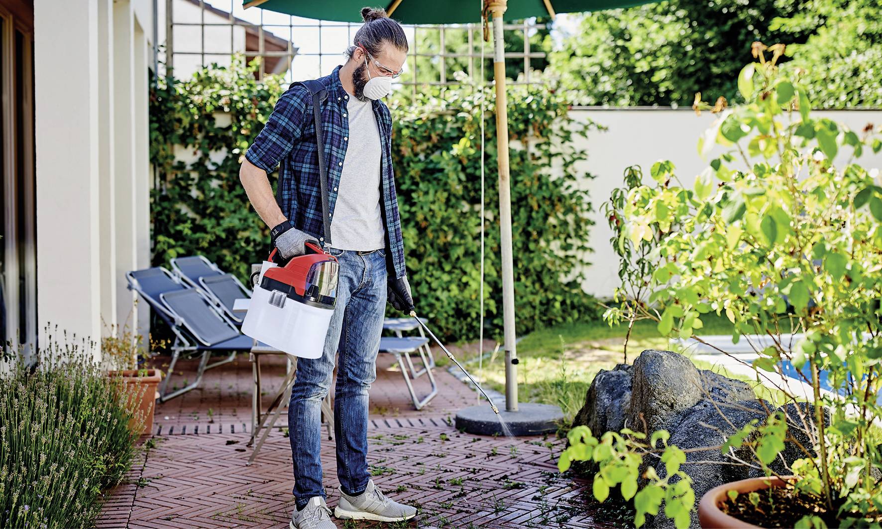 Een persoon met een beschermmasker en handschoenen sproeit planten in de tuin. Op de achtergrond zijn tuinmeubels en groene planten te zien.