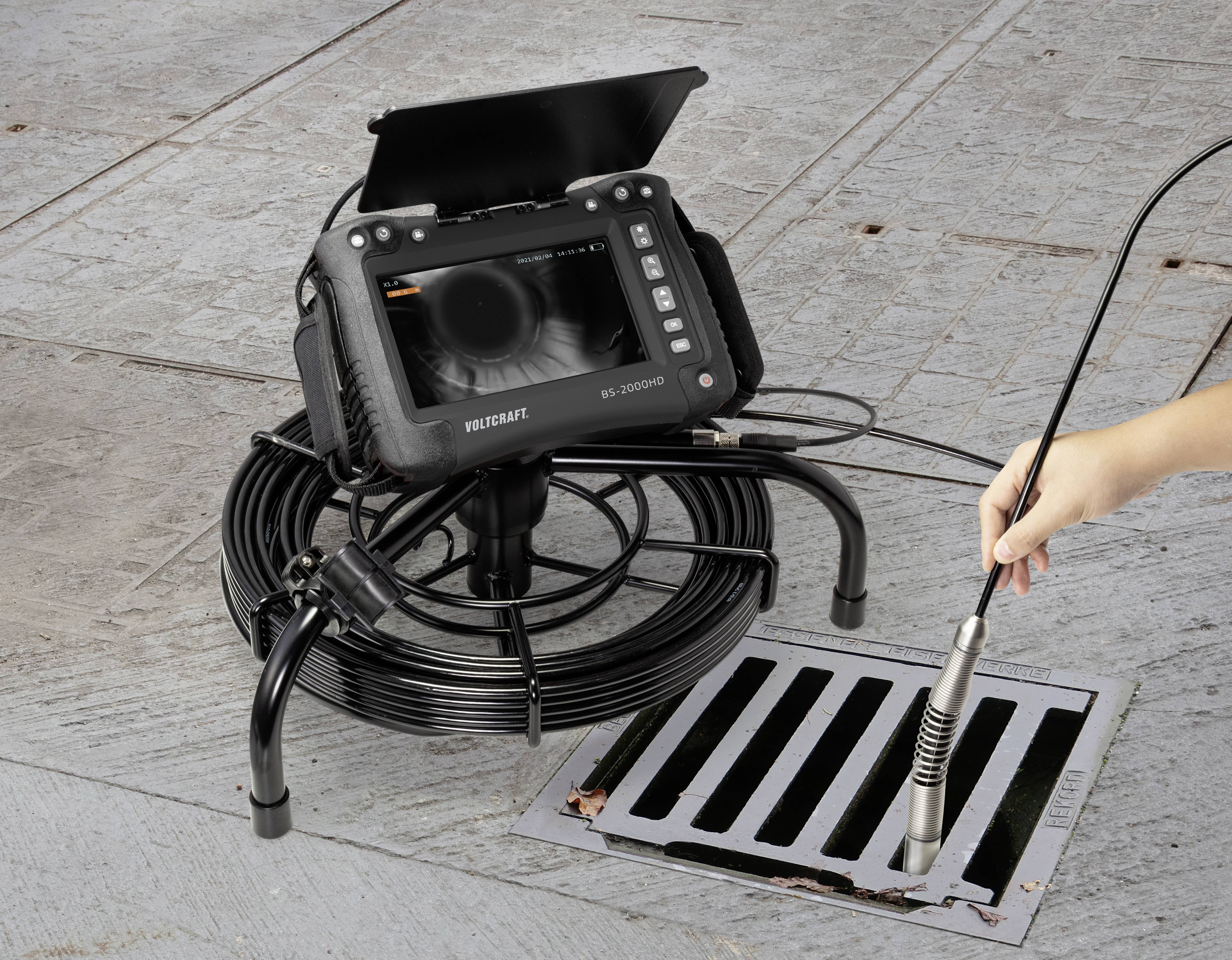 Een hand brengt een camera met een flexibel kabel in een straatriool, terwijl een bewakingsmonitor het binnenste weergeeft.