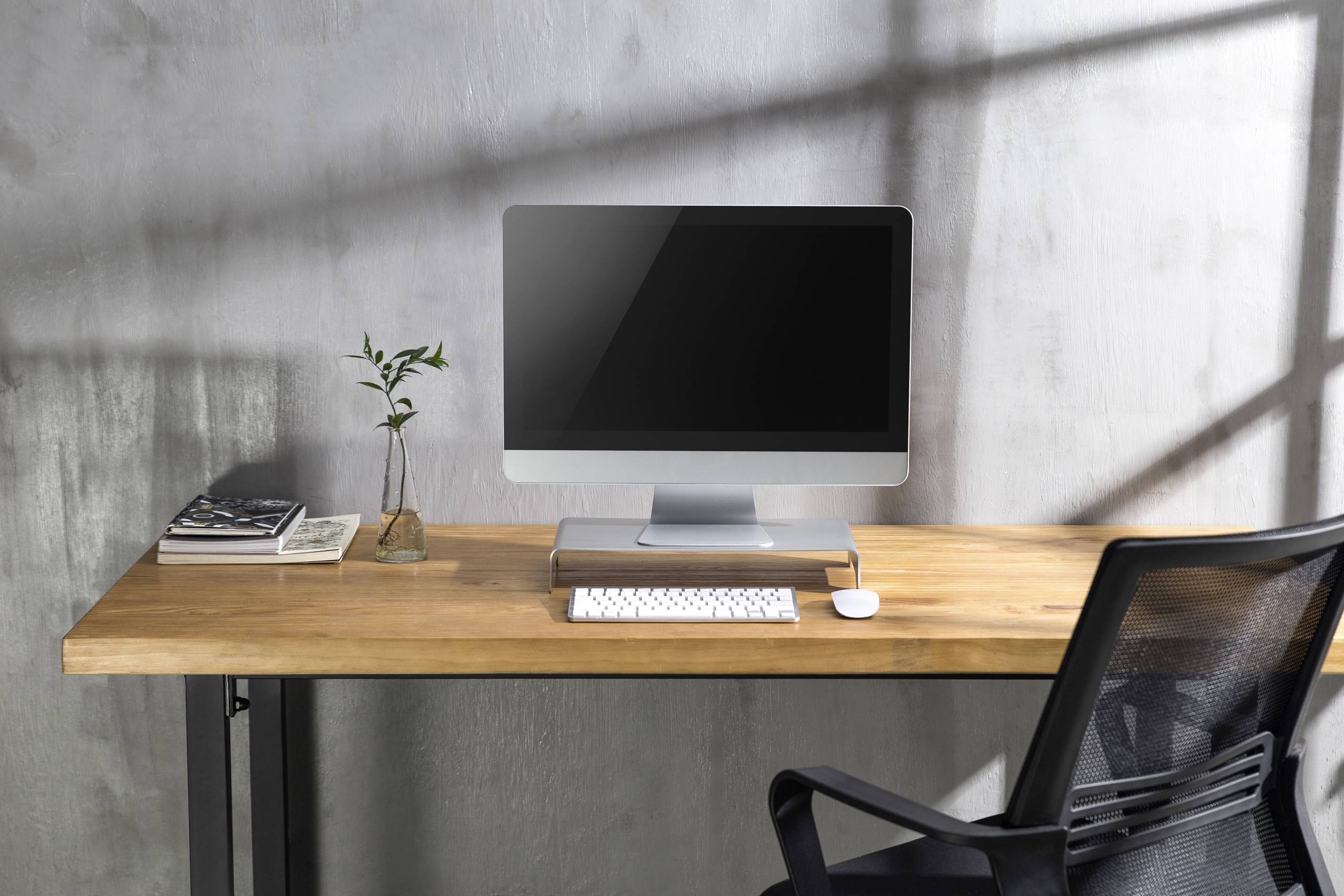 Een moderne bureau met computer, toetsenbord, muis, een stapel boeken en een kleine vaas met een plant. Op de achtergrond een grijze muur.