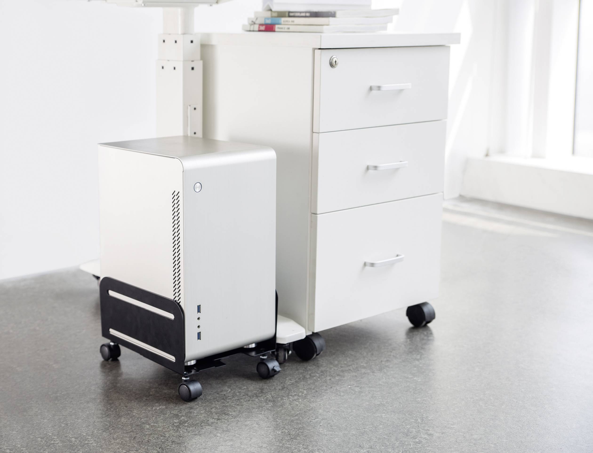 Een zilveren computer op een standaard staat naast een witte rolcontainer met drie laden op een betonnen vloer in een lichte ruimte.