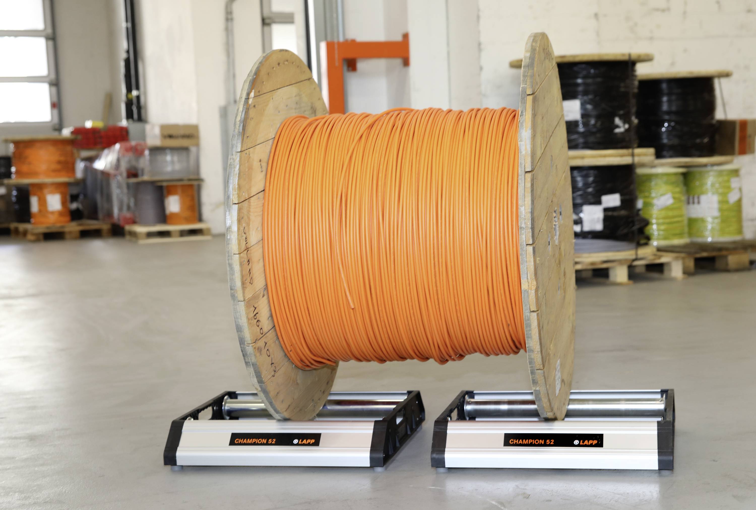 Een grote kabel haspel met oranje kabel staat in een opslagruimte. Op de achtergrond zijn nog meer kabel haspels op pallets zichtbaar.