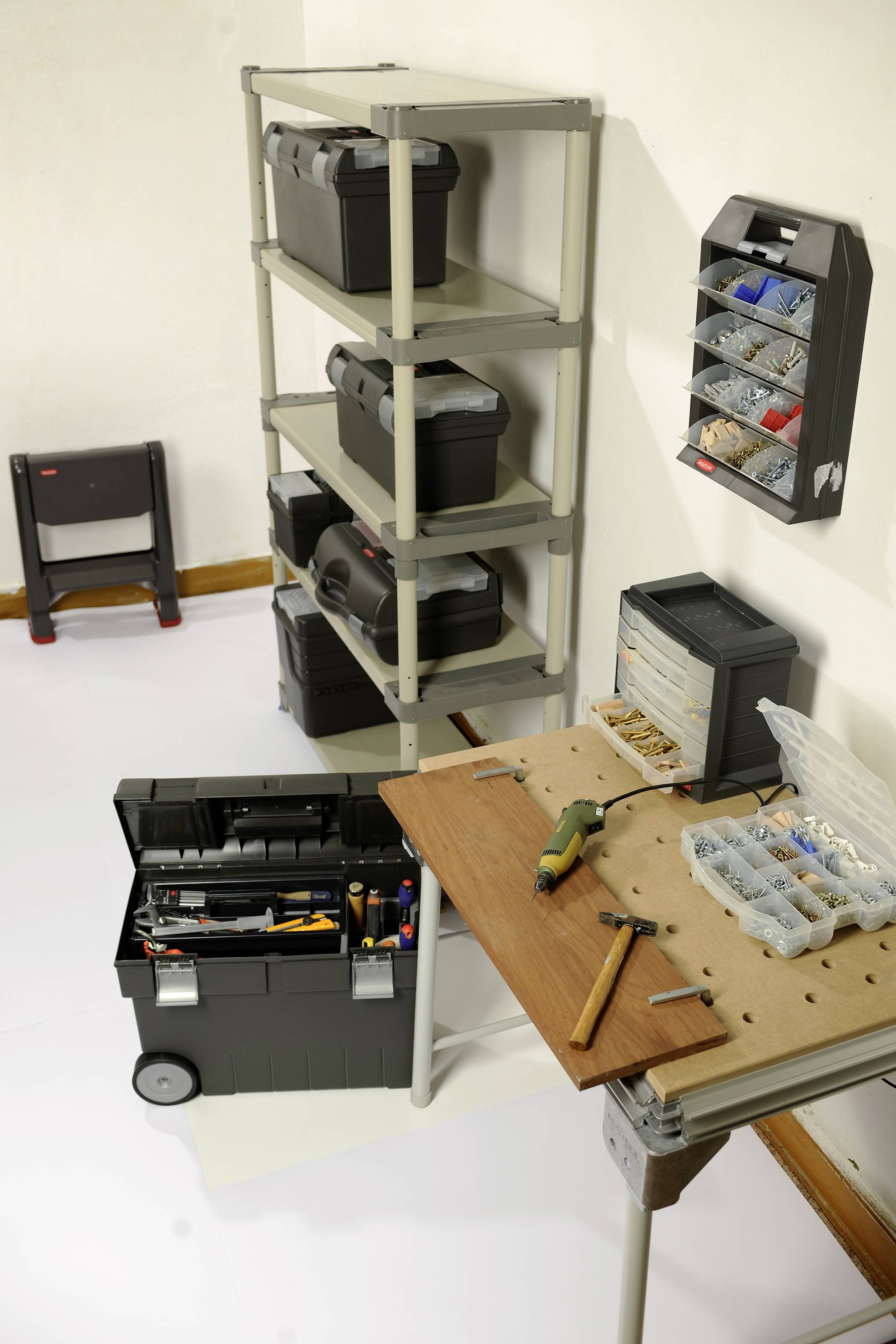 Werkplaats met rekken vol gereedschapskisten, wandhouders voor kleine onderdelen, en een werktafel met gereedschappen zoals hamer en boormachine.
