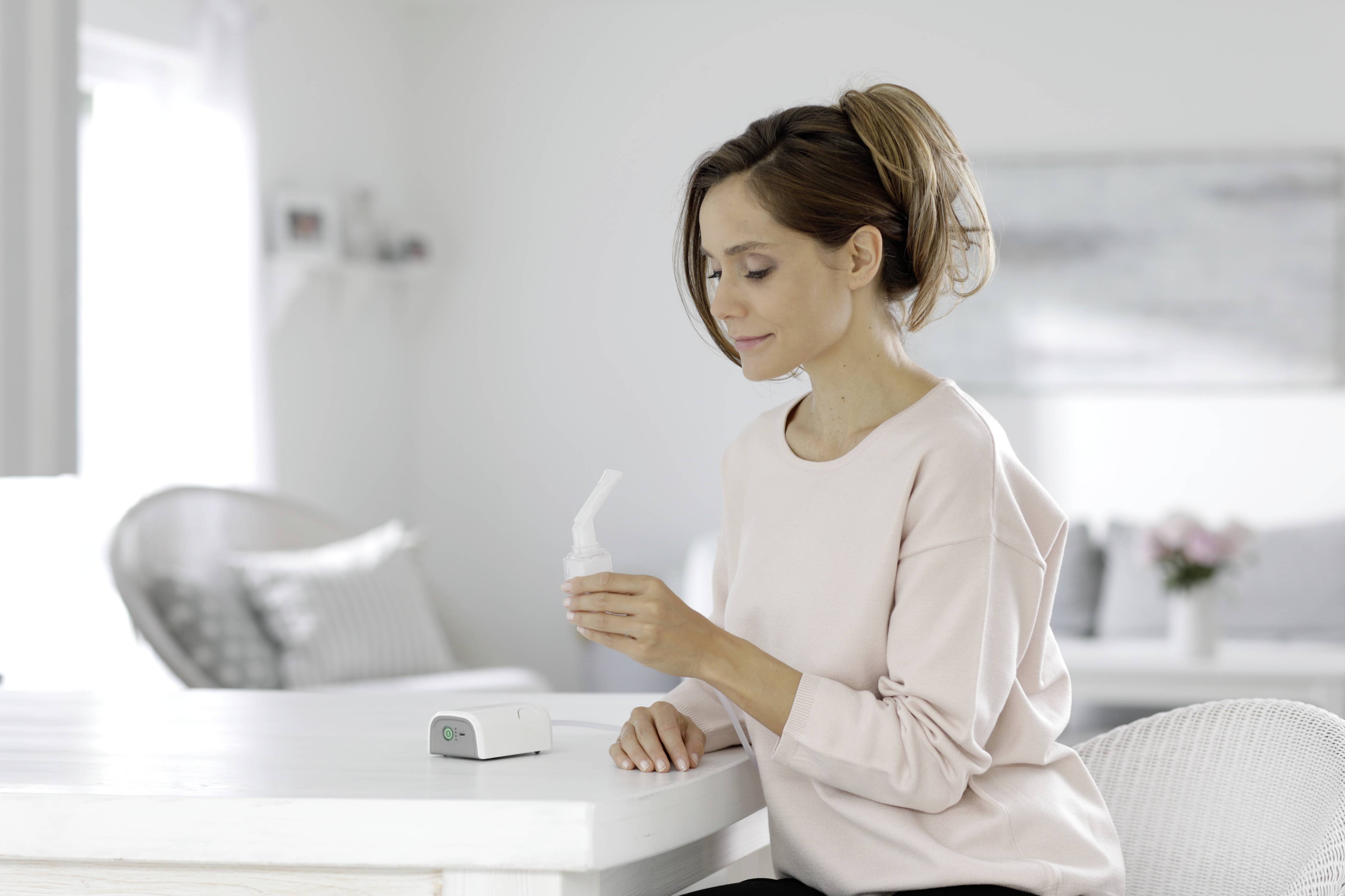 Een vrouw zit aan een tafel en houdt een inhalatieapparaat vast. Op de achtergrond zijn een lichte woonkamer en een bank zichtbaar.