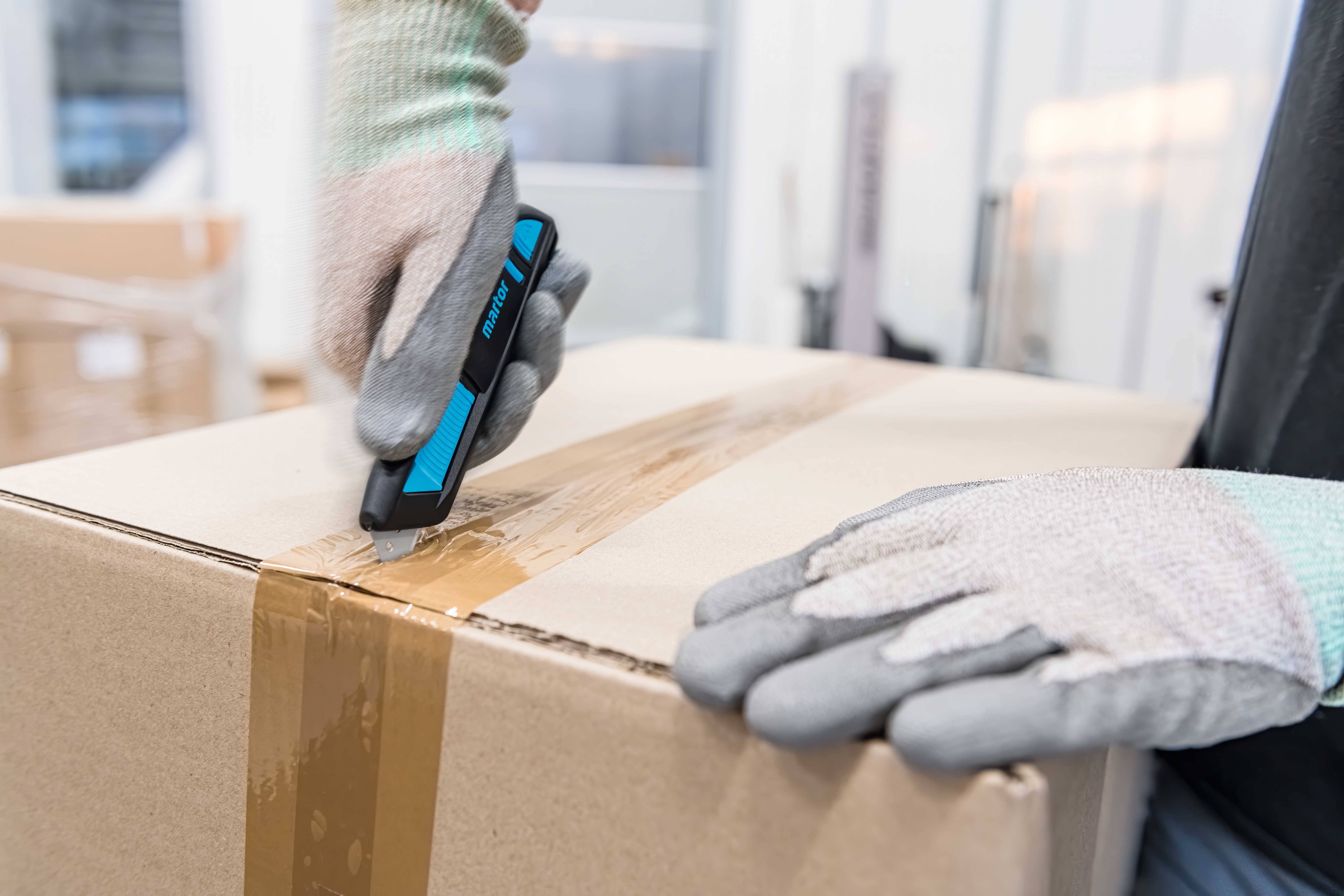 Een persoon opent een kartonnen doos met een blauw stanleymes, beschermd door grijze handschoenen. Op de achtergrond bevinden zich onscherpe opslagrekken.