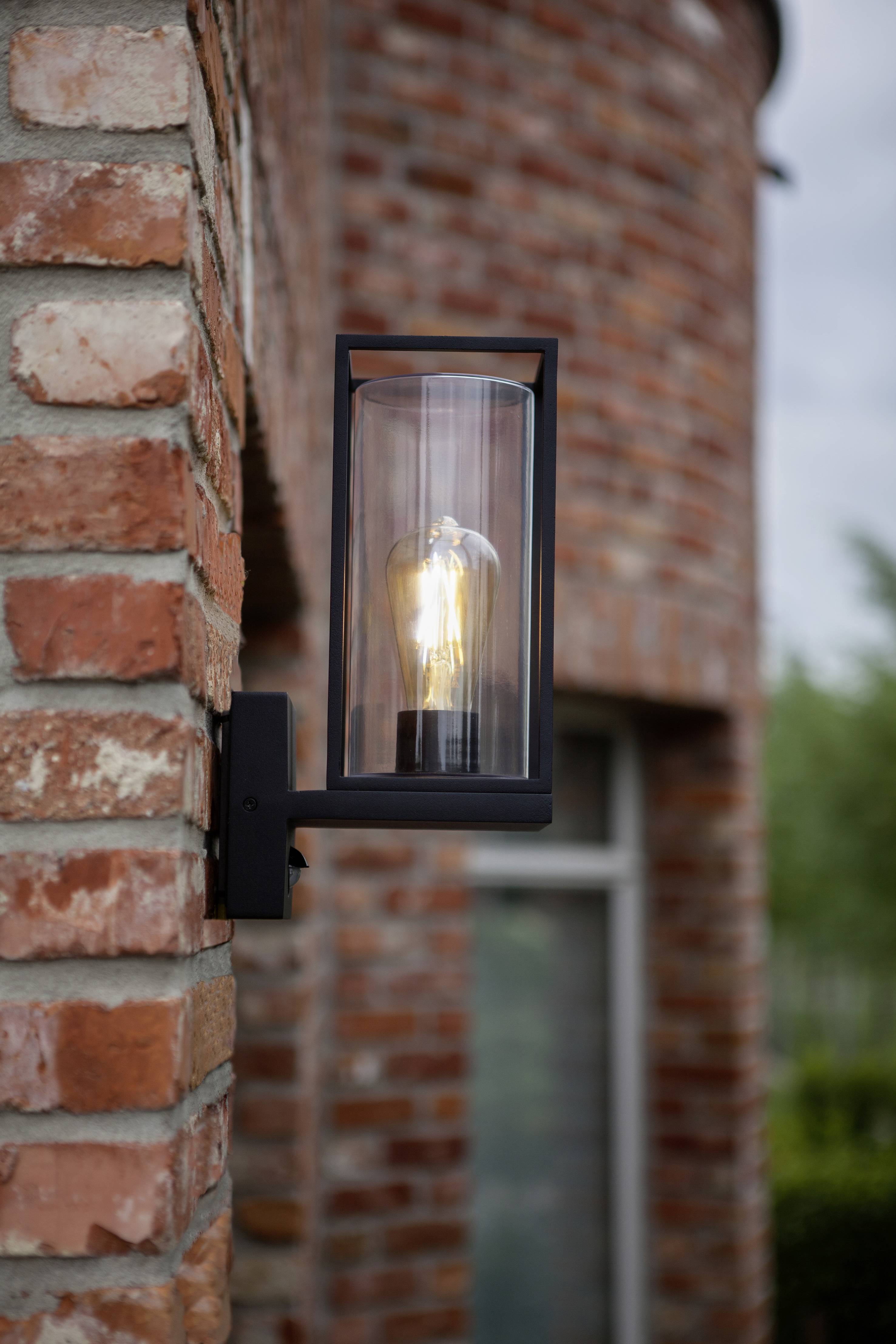 'Buitenlamp gemonteerd aan een bakstenen muur, ingeschakeld, met op de achtergrond wazig een raam zichtbaar.'