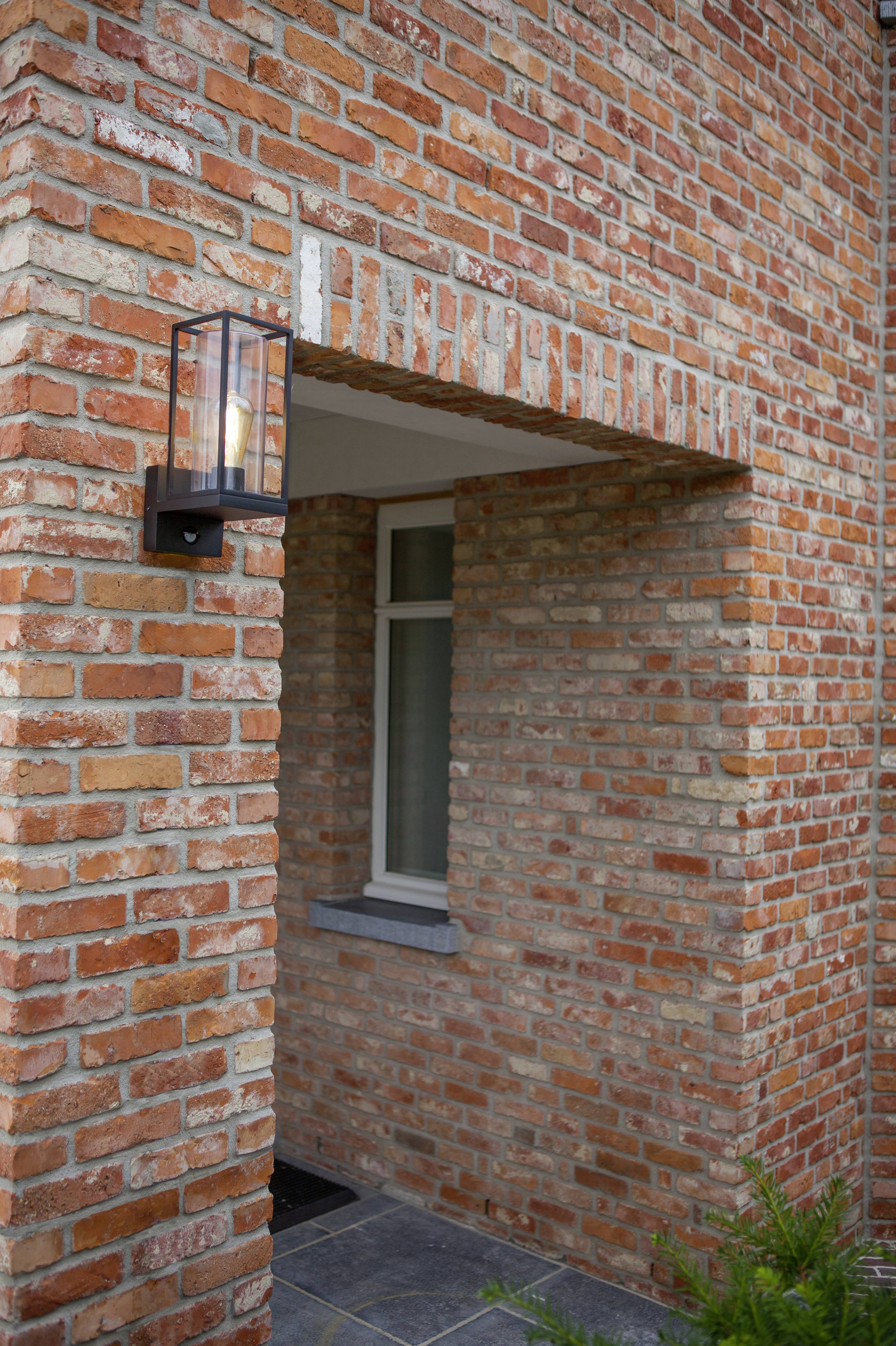 Bakstenen gevel van een huis met een kleine ingangspartij. Aan de muur hangt een zwarte lantaarn.