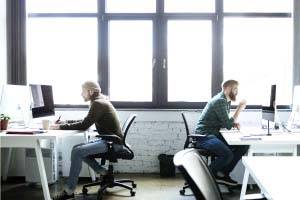 Twee personen werken aan computers in een modern kantoor met grote ramen. De sfeer is geconcentreerd en productief.