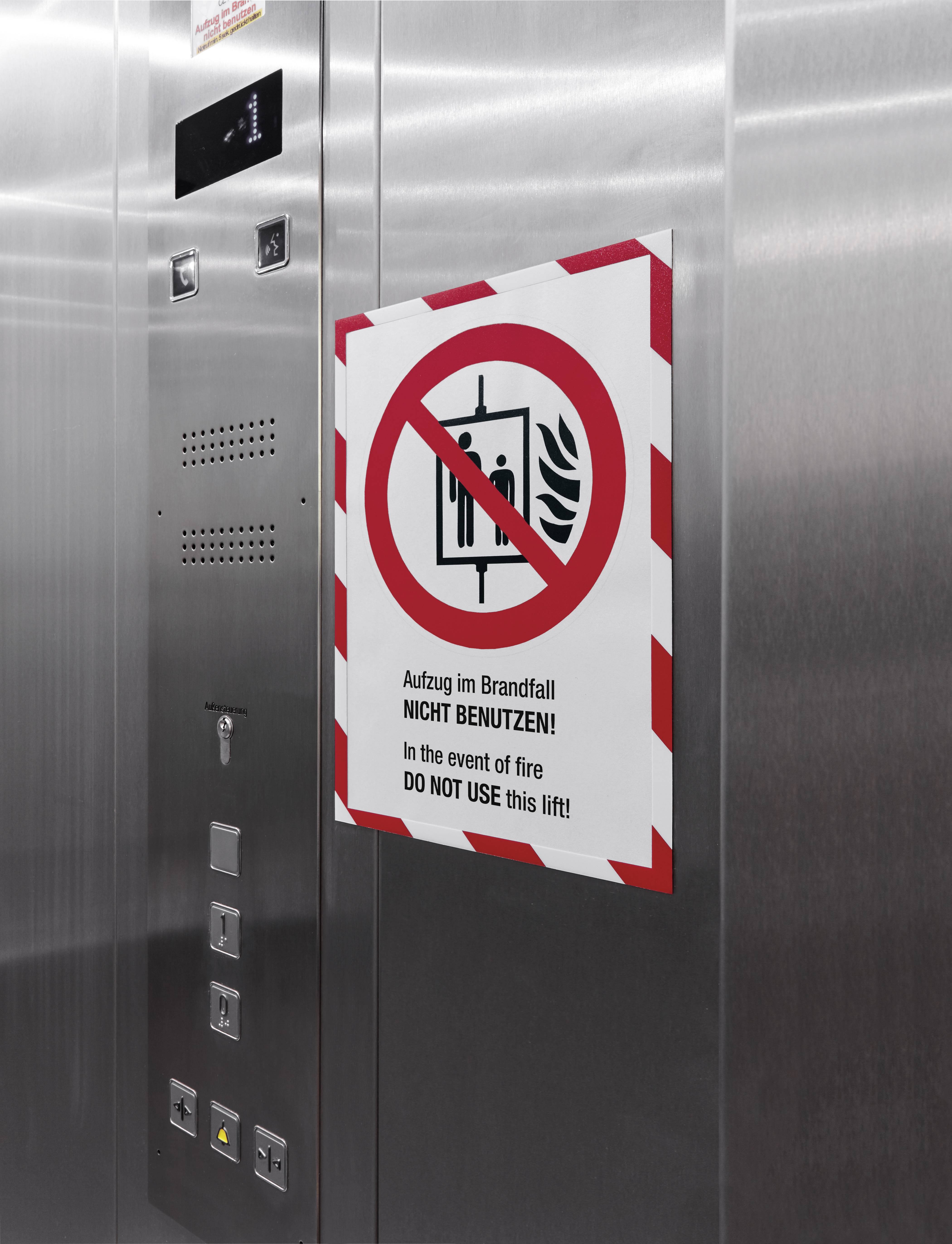 'Niet gebruiken'-bord in de lift: 'Lift in geval van brand NIET GEBRUIKEN', rood verbodssymbool, roestvrij stalen wand op de achtergrond.