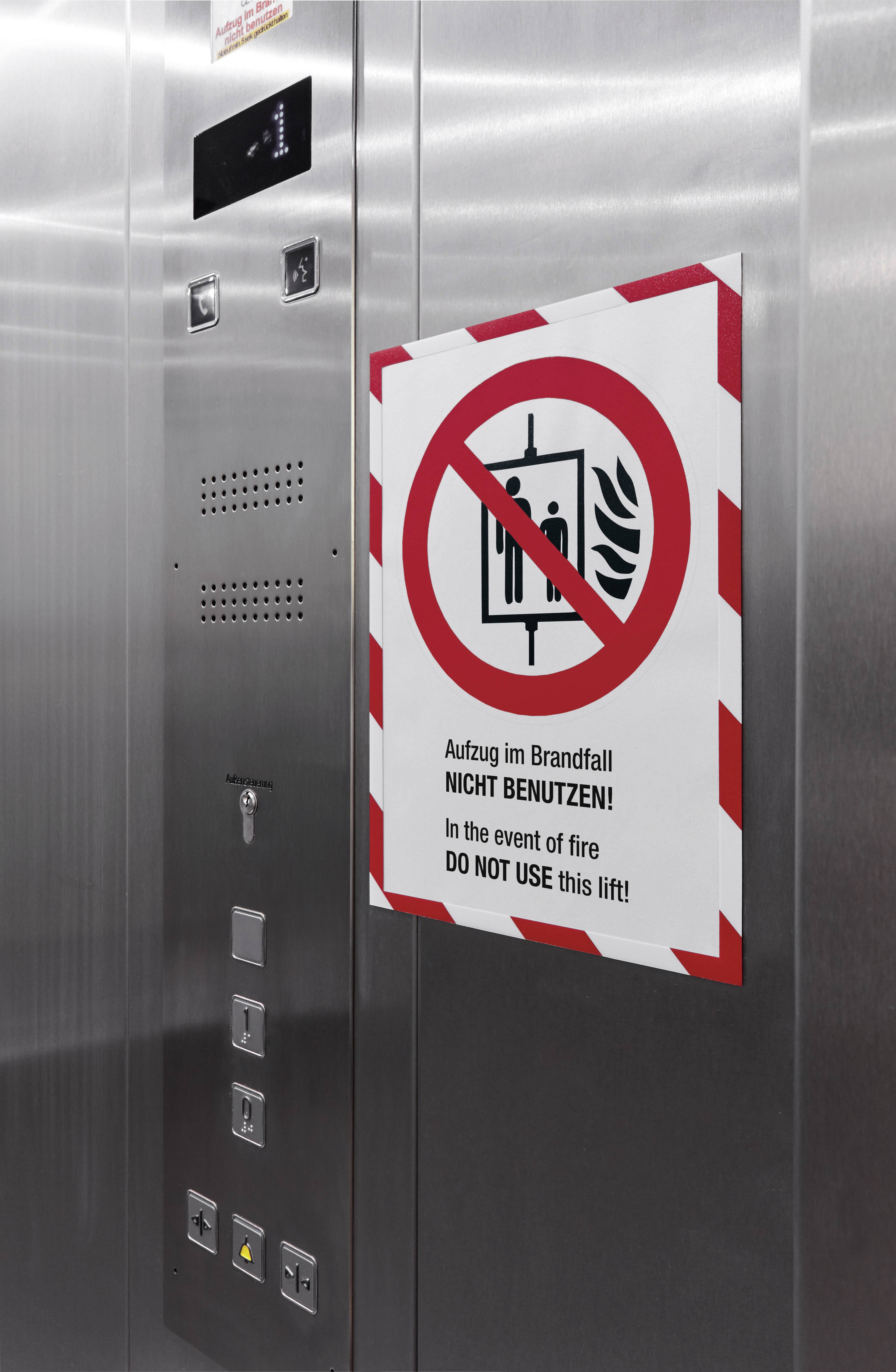 Een sticker in de lift waarschuwt: 'Lift niet gebruiken bij brand'. Het bord toont een doorgestreept liftsymbool met vlammen.