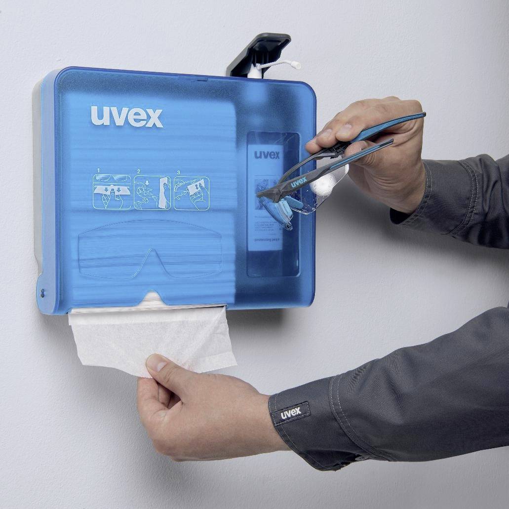 'Een persoon haalt een veiligheidsbril uit een blauwe uvex-dispenser aan de muur. Een andere hand houdt een doek vast.'
