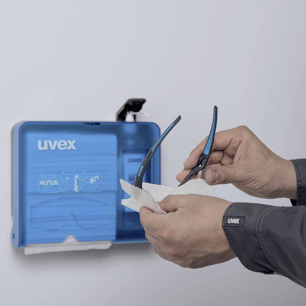 Een persoon reinigt een veiligheidsbril met een doek, voor een wanddispenser voor reinigingsmiddelen met 'uvex' opschrift.