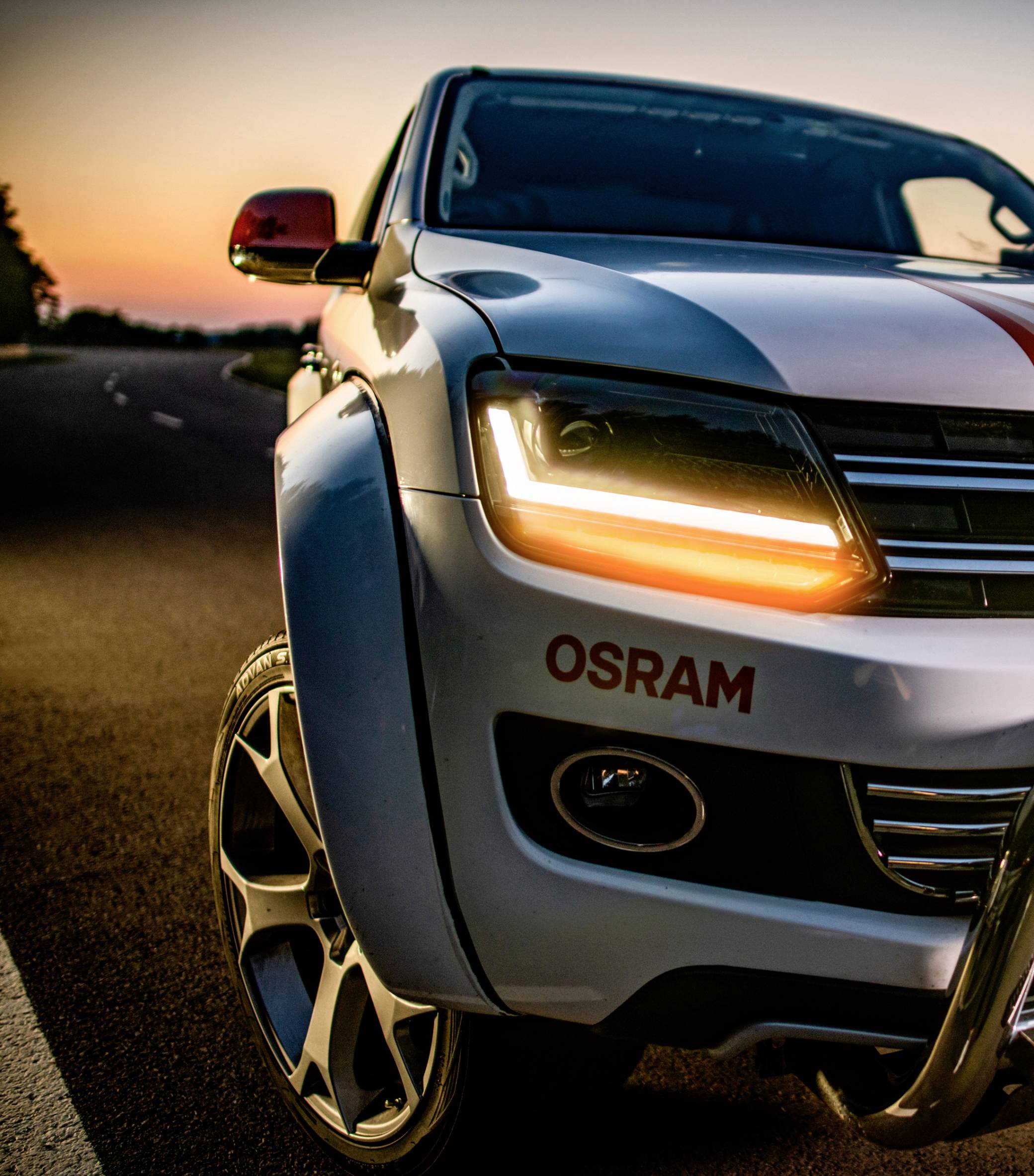 Een witte SUV met ingeschakelde koplampen staat op een weg bij zonsondergang. "OSRAM" staat op het voertuig geschreven.