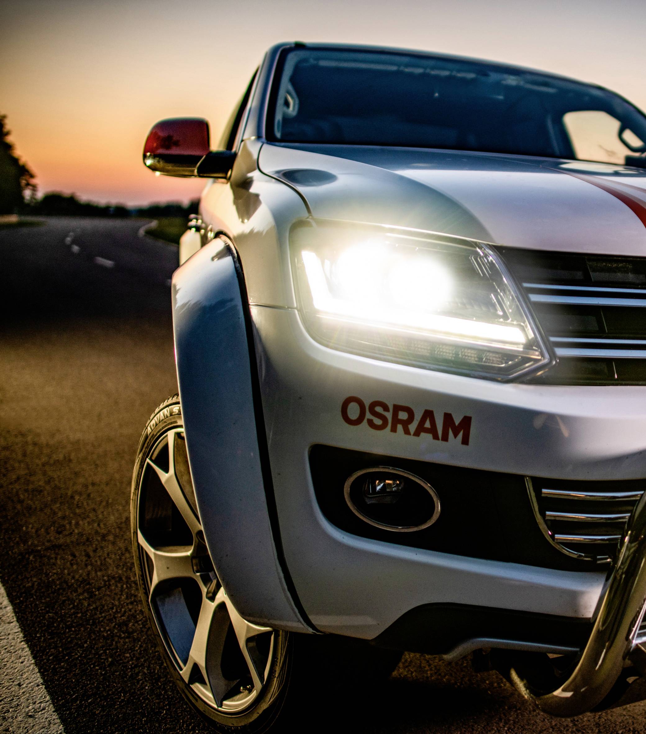 Terreinwagen op een weg bij zonsondergang met ingeschakelde koplampen. 'OSRAM' op de voorkant vermeld.