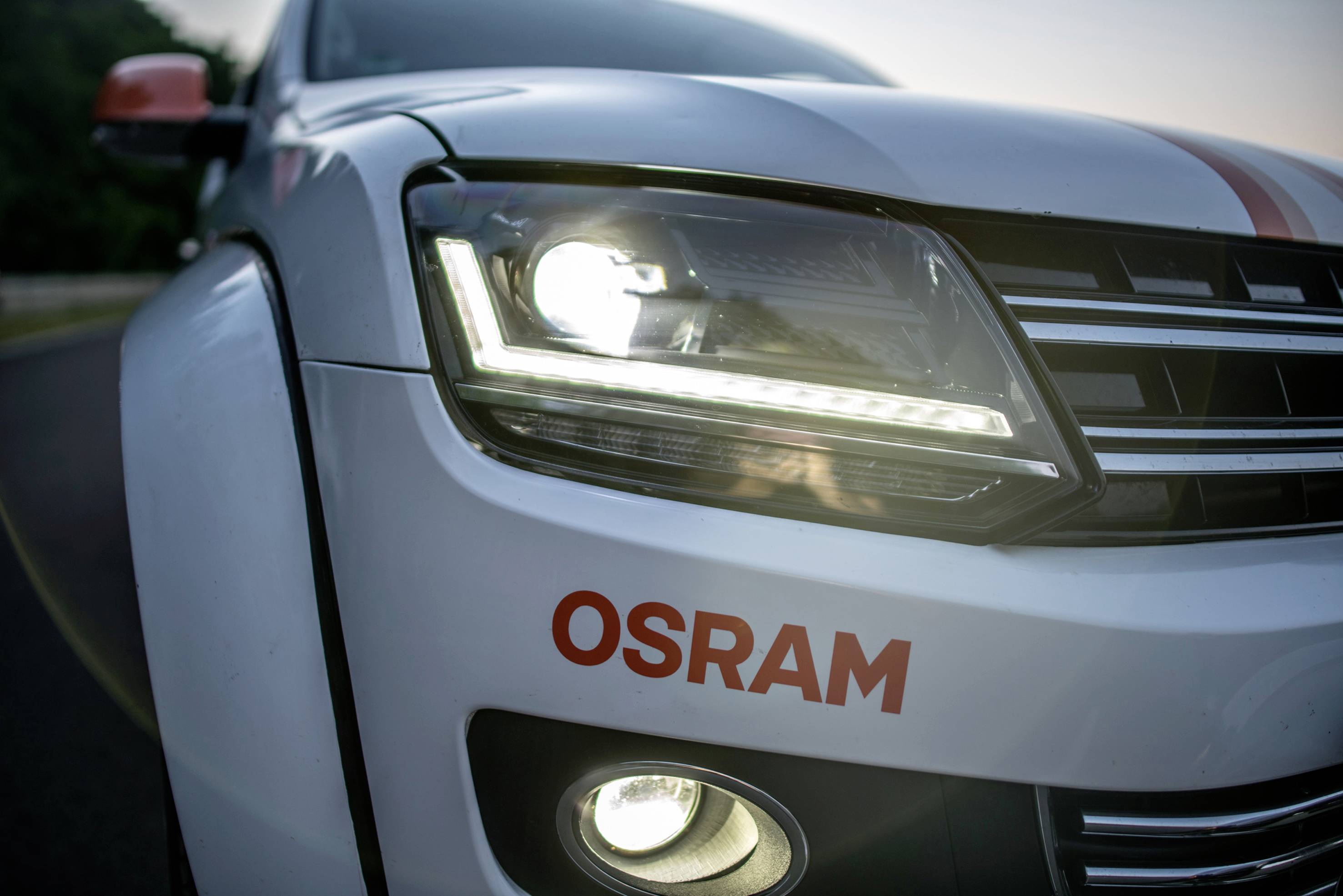 Witte auto met OSRAM-opschrift, hoofdkoplamp en mistlamp schijnen op een weg.