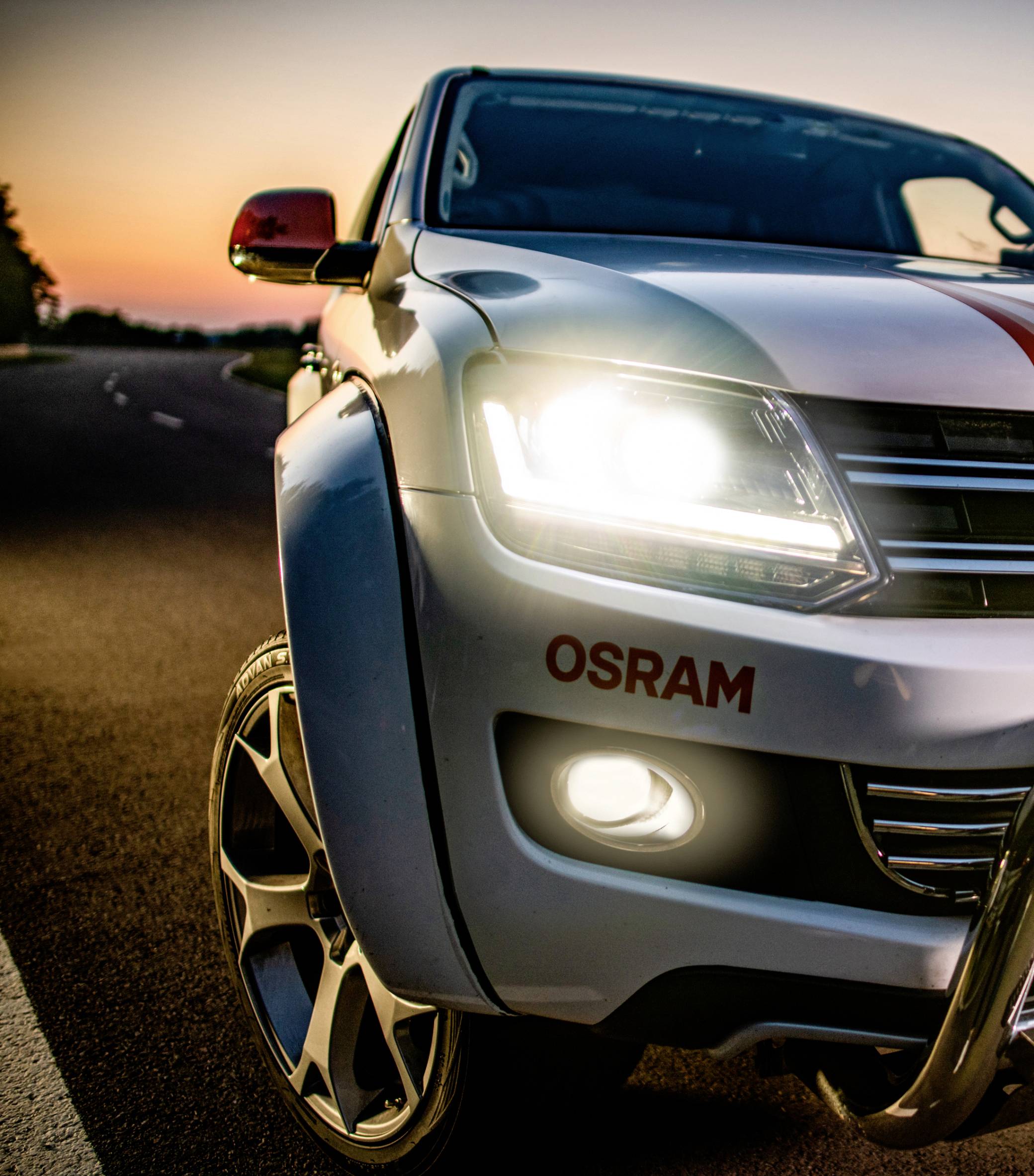 Close-up van een auto met ingeschakelde koplampen op een weg tijdens zonsondergang. De merknaam 'OSRAM' is zichtbaar.