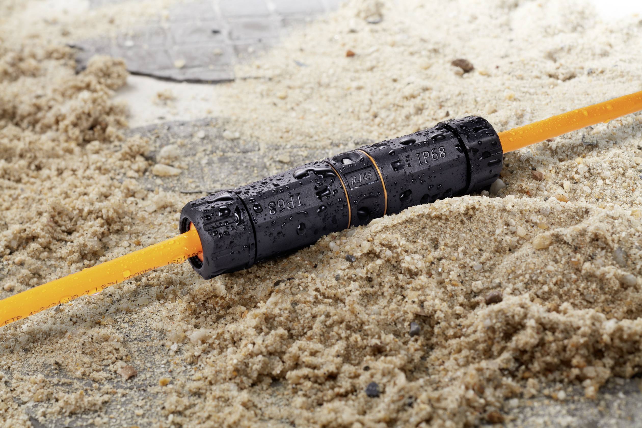 Een waterdichte zwarte connector met een oranje leiding rust op een zandondergrond.