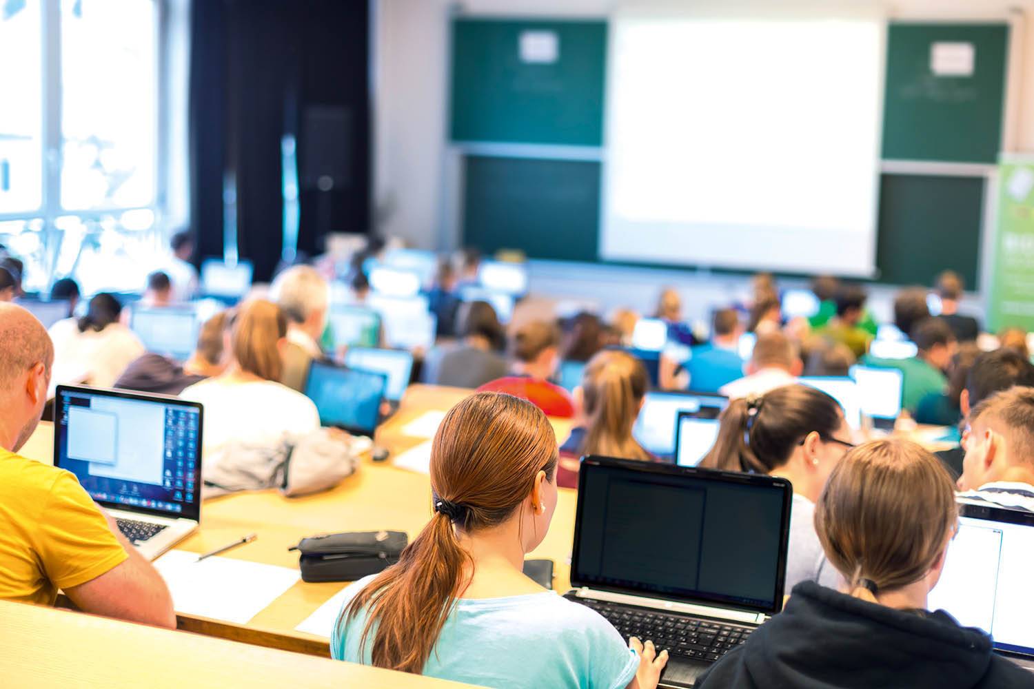 Studenten zitten in een collegezaal, werken op laptops en concentreren zich op een presentatie aan een bord.
