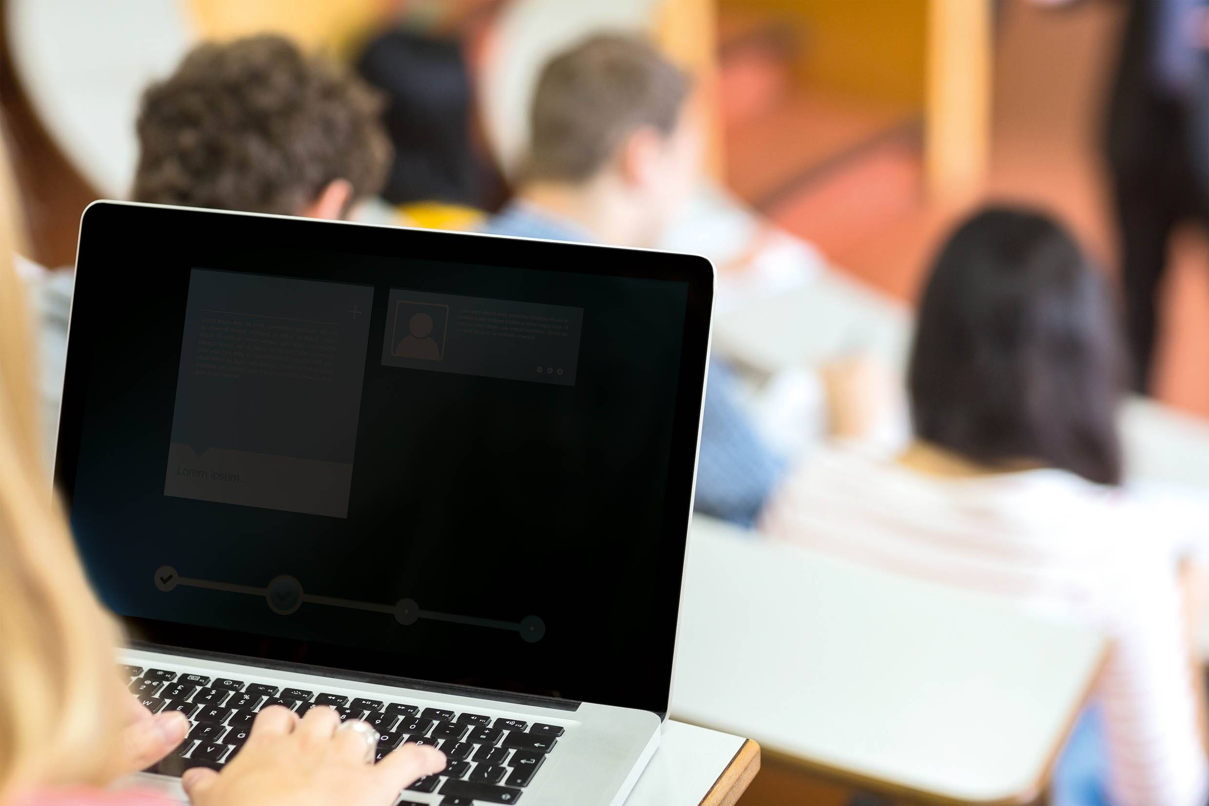 Een persoon typt op een laptop in een klaslokaal. Op de achtergrond zijn onscherpe studenten te zien die een lezing volgen.