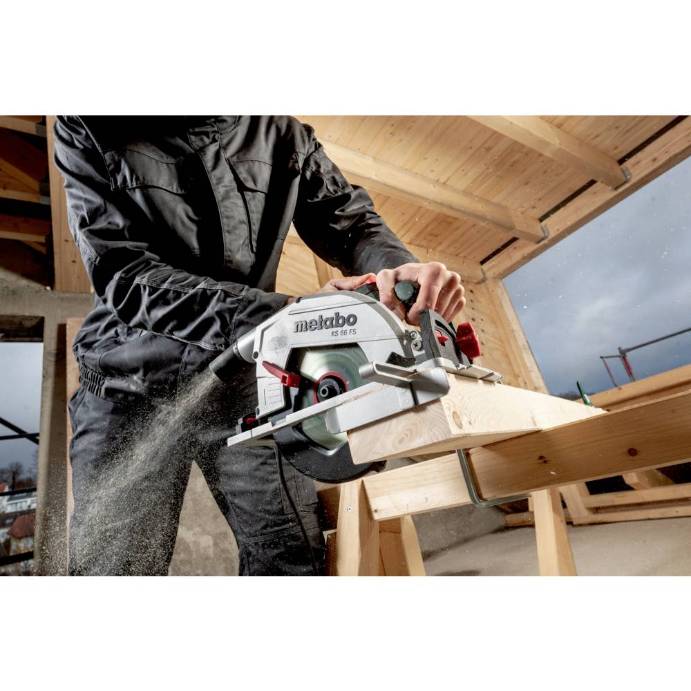 Een persoon gebruikt een Metabo cirkelzaag om door een houten balk te zagen in een gedeeltelijk geconstrueerd gebouw met zichtbare houten constructiebalken.