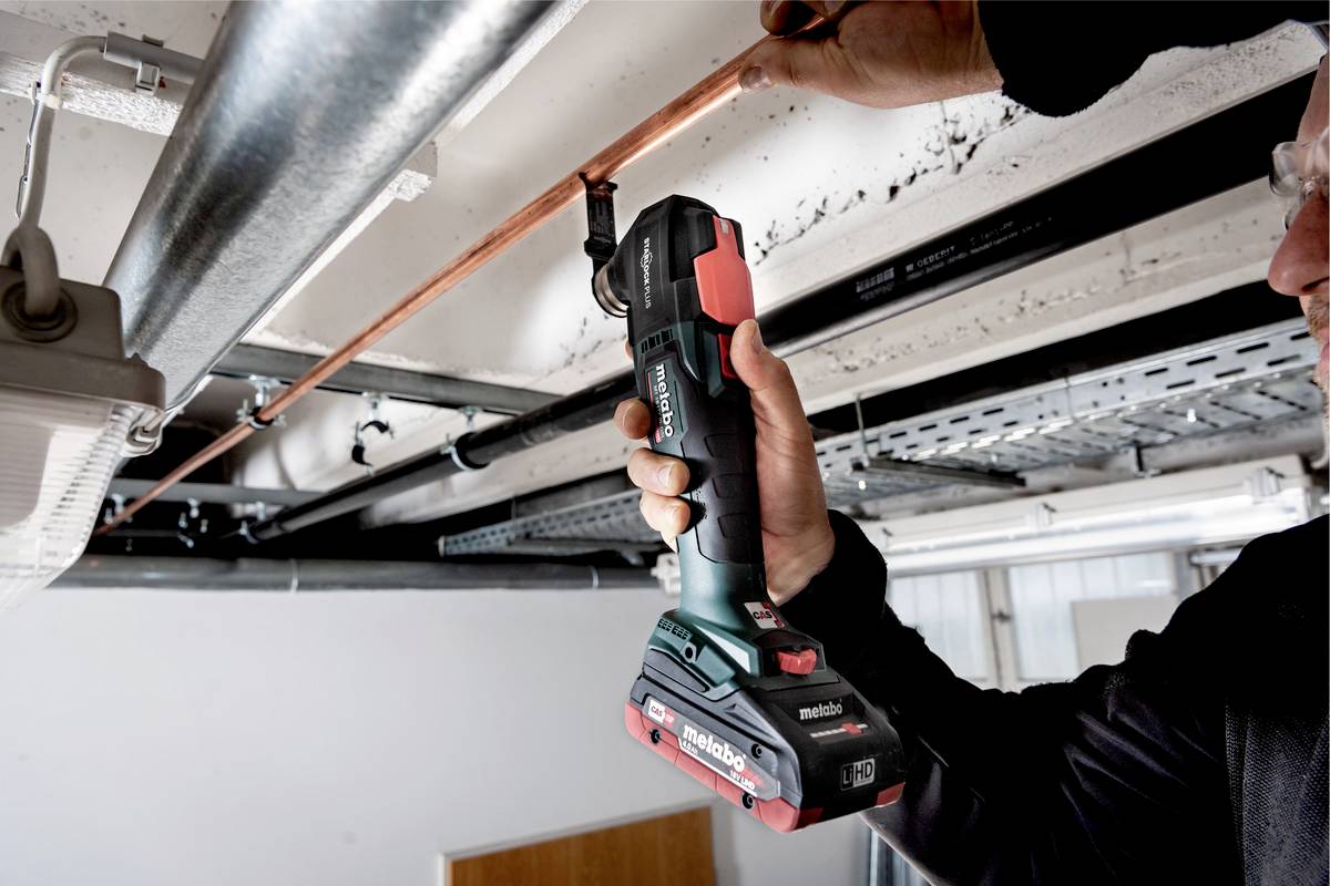 Een persoon gebruikt een elektrisch gereedschap om een koperen buis aan het plafond te bevestigen. Het gaat om een accugereedschap.