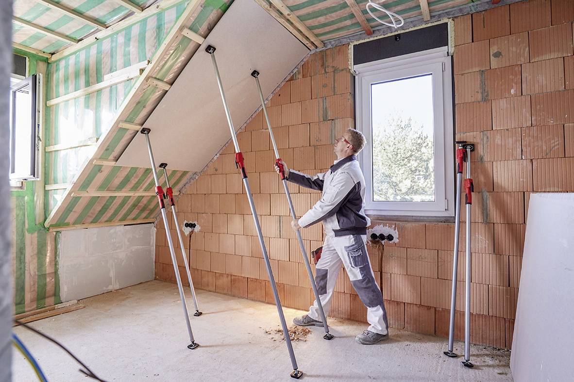 Een persoon installeert gipsplaten aan het plafond van een nog onafgewerkte zolder. Er zijn steunen aanwezig ter ondersteuning.