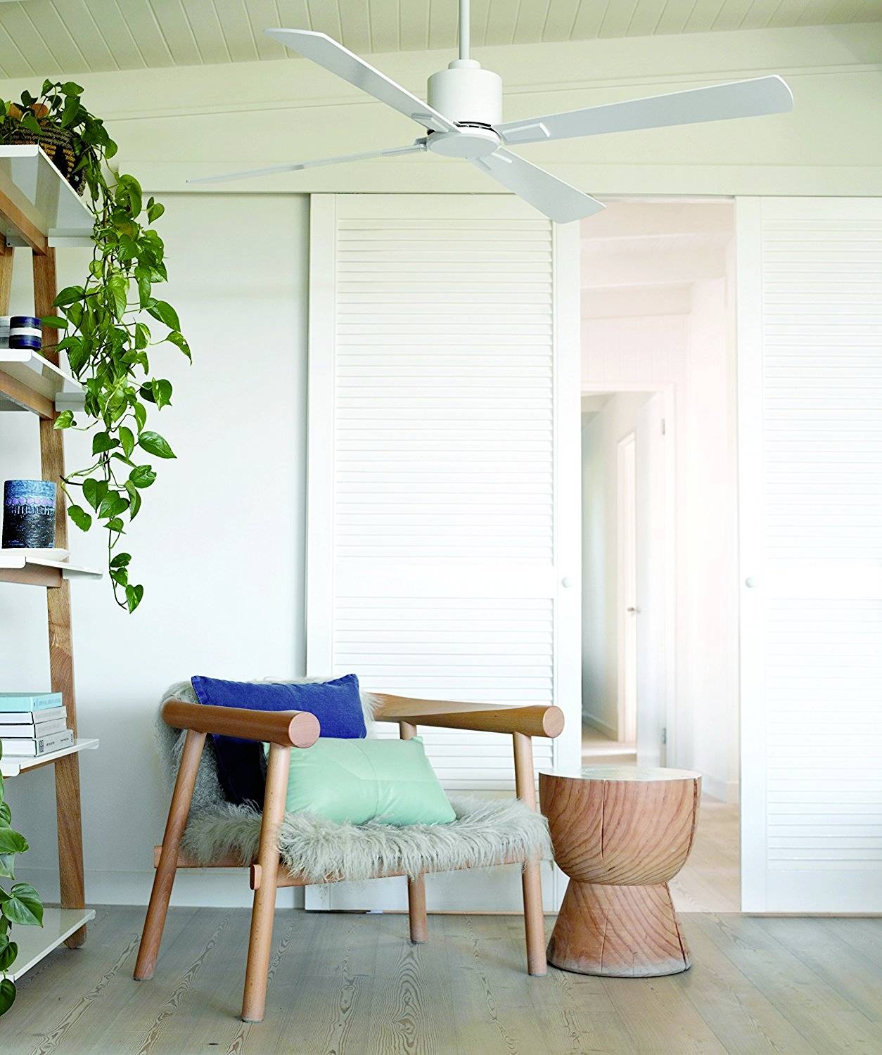 Lichte woonkamer met een fauteuil, decoratieve kussens, een houten krukje, een rek met planten en een plafondventilator.