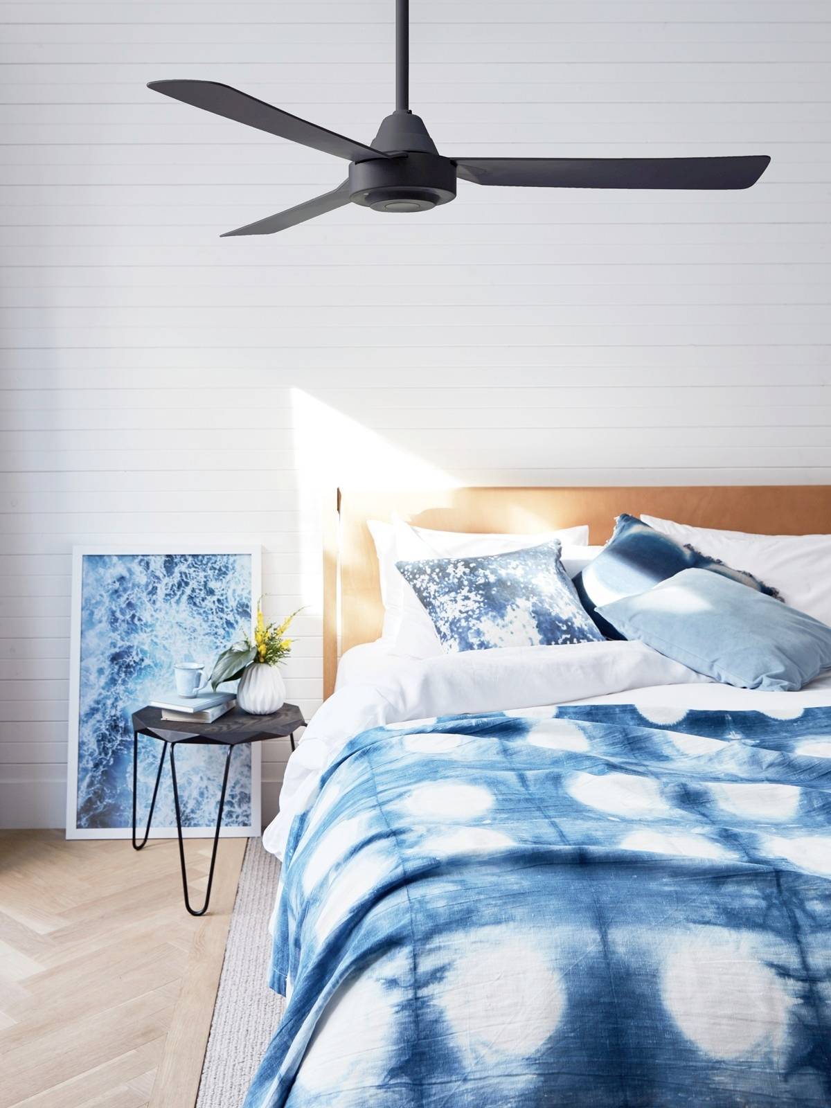 Een modern slaapkamer met een bed dat blauwe batik beddengoed en witte kussens heeft. Naast het bed staat een kleine tafel met een vaas.