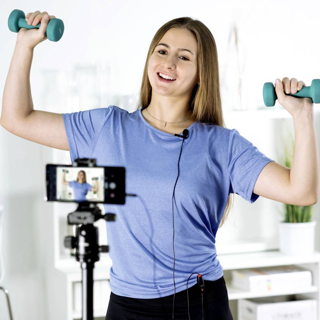 Een vrouw in sportkleding tilt halters voor een camera terwijl ze praat. Ze lijkt een fitnessvideo op te nemen.