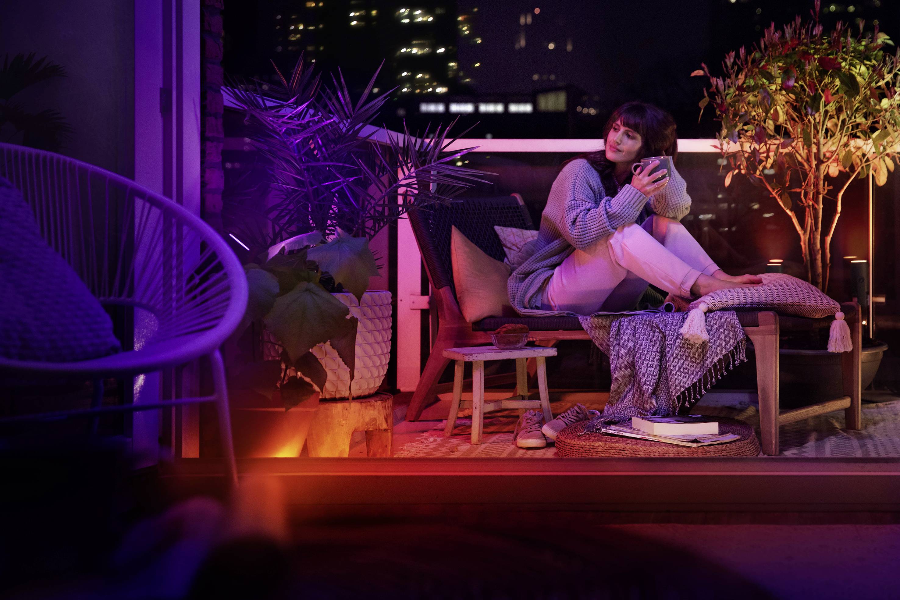 Een vrouw zit ontspannen op een balkon 's avonds, omringd door planten en zacht licht, en drinkt uit een kopje.