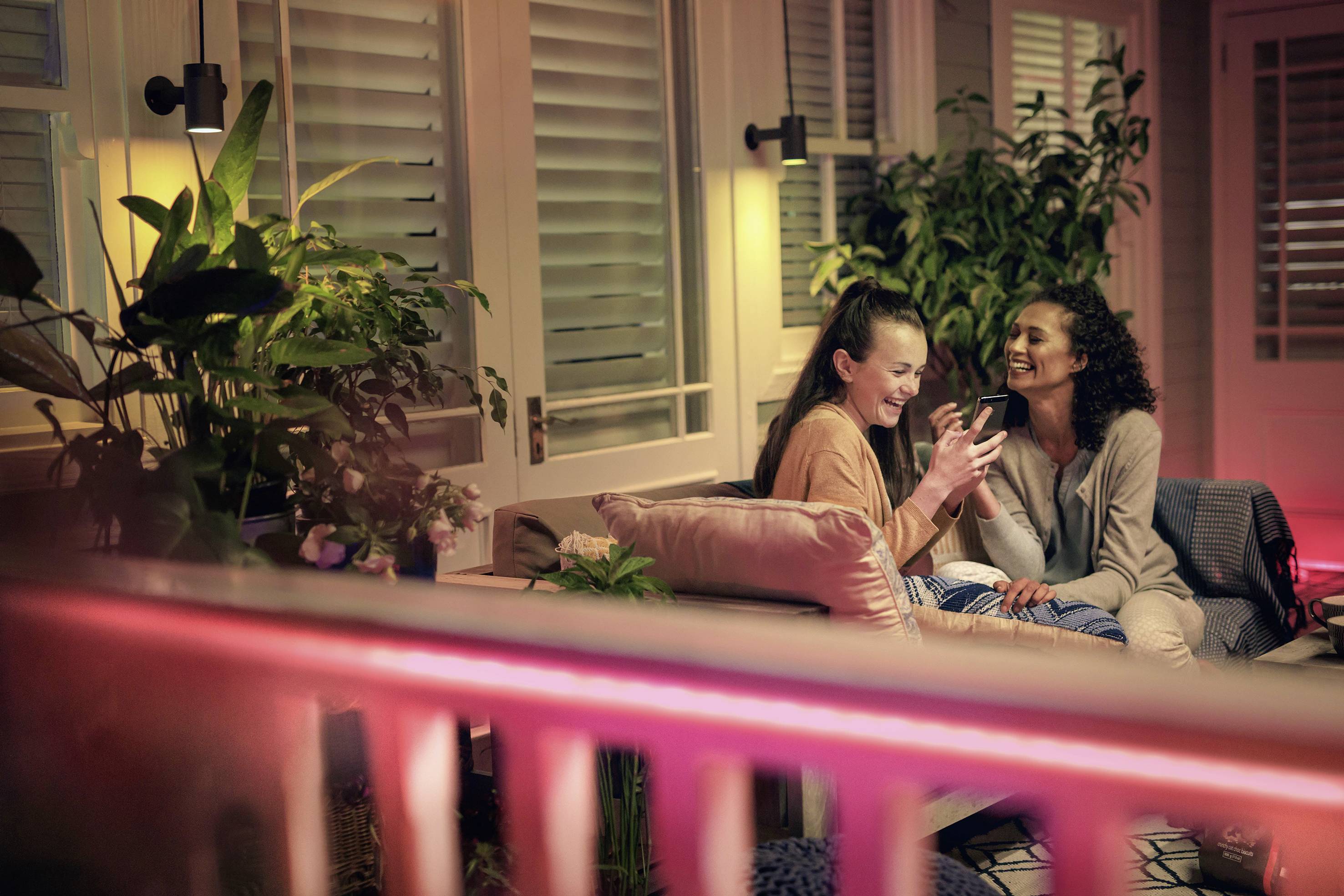 Twee personen zitten lachend op een terras vol planten en kijken samen naar een smartphone. Ze lijken ontspannen en gelukkig.