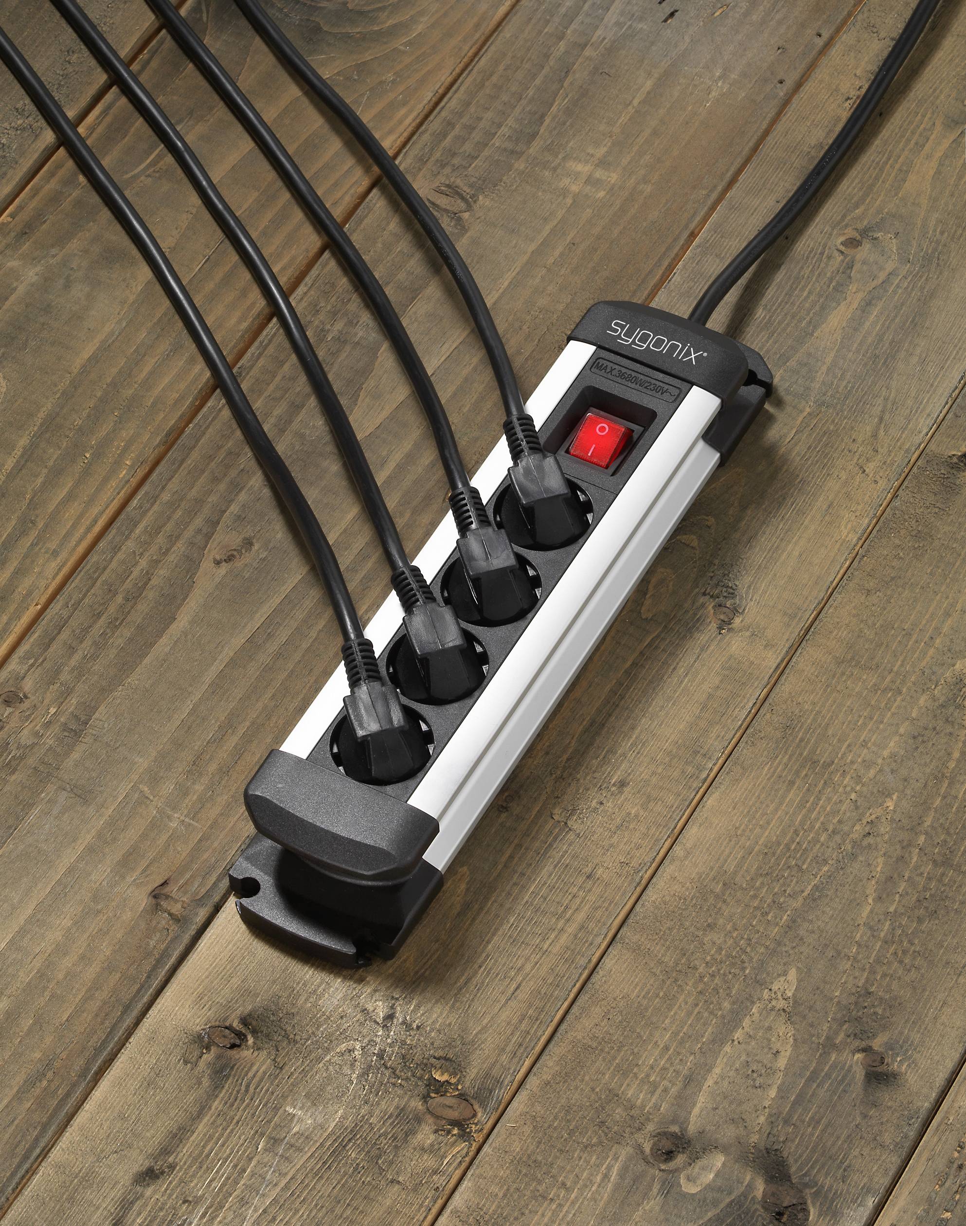 Een zwarte stekkerdoos met vijf aangesloten kabels, geplaatst op een houten vloer. Een rode netschakelaar is zichtbaar.