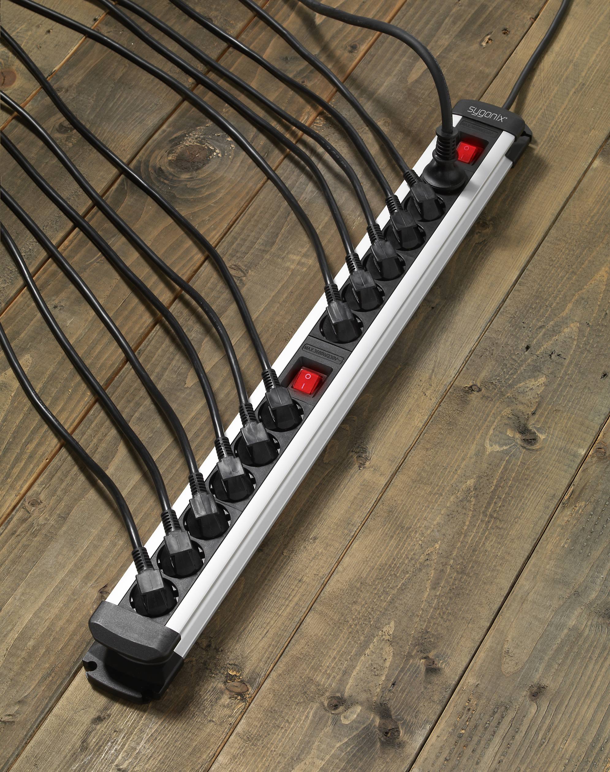 Een lange stekkerdoos met twaalf aangesloten zwarte kabels ligt op een houten vloer. Twee rode schakelaars zijn zichtbaar.