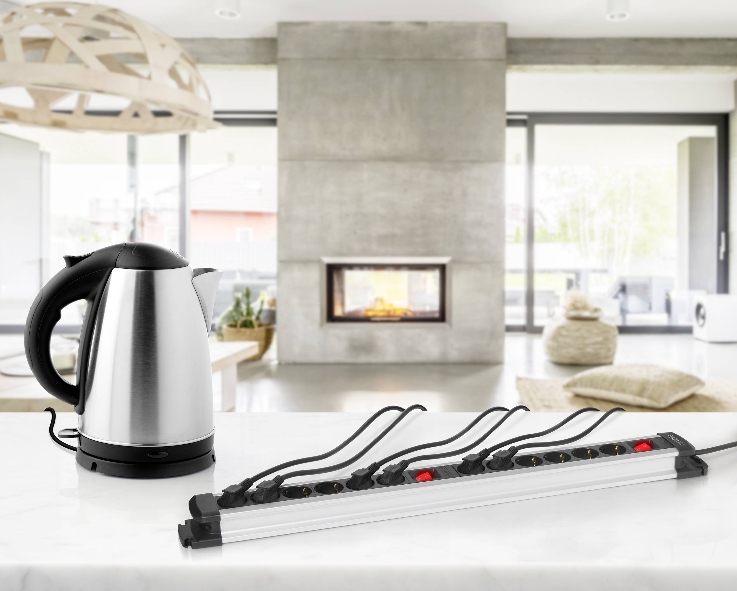 Een waterkoker staat op een aanrecht; naast hem ligt een lange stekkerdoos. Op de achtergrond is een moderne woonkamer met een open haard zichtbaar.