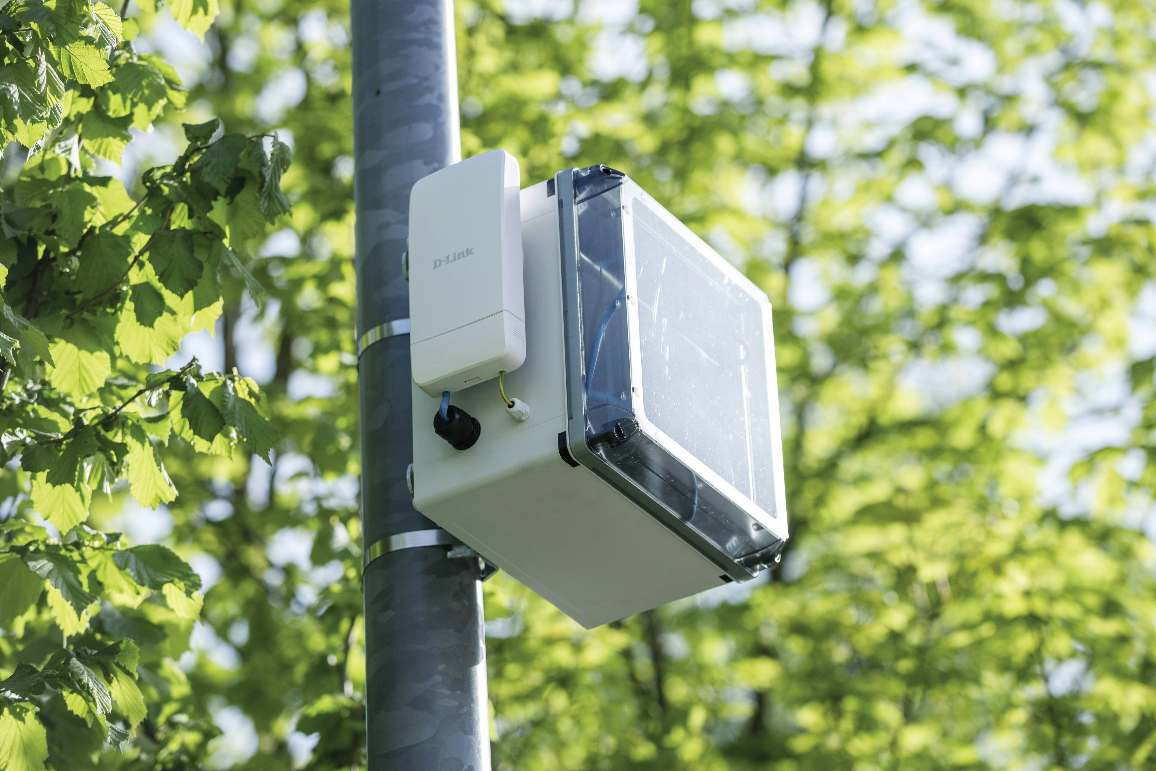 Een weerstation met een sensor en een zonnepaneelbehuizing is bevestigd aan een mast. Achtergrond: groene bladeren.