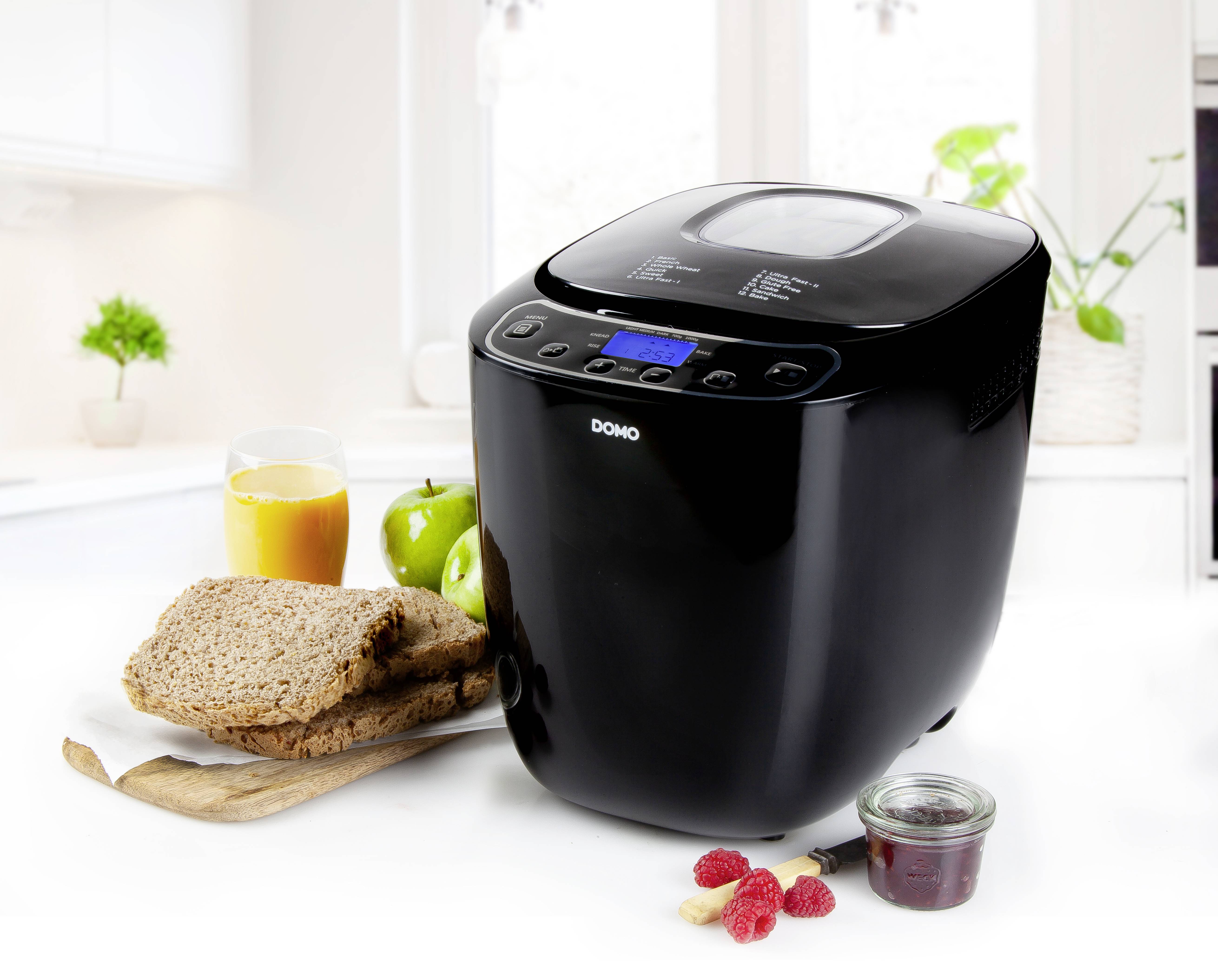 Broodbakmachine in moderne keuken, omringd door volkorenbrood, een glas sinaasappelsap, appels, jam en bessen.