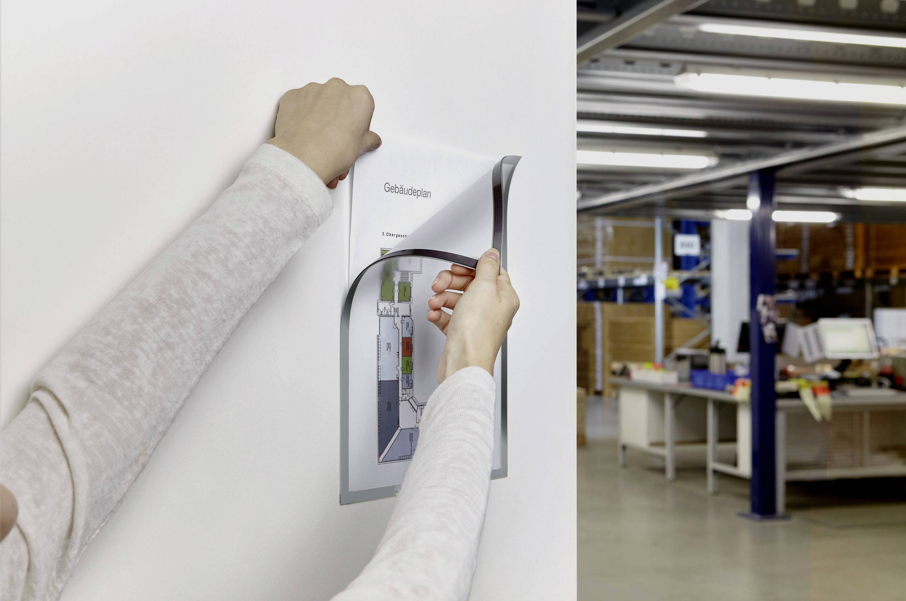 Een persoon hangt een informatieblad met de titel 'Gebouwplattegrond' aan een muur. Op de achtergrond zijn kantoormeubels zichtbaar.