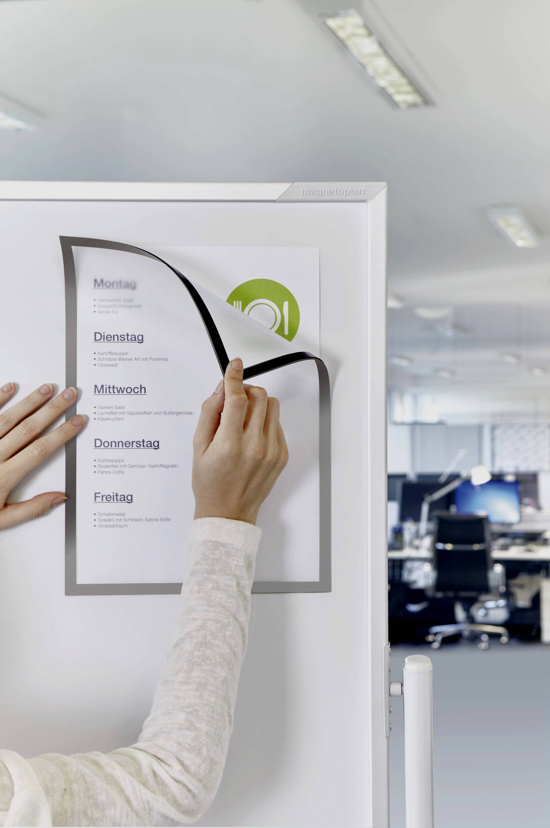 Een persoon vervangt een blad op een whiteboard-kalender in een modern kantoor. De dagen van de week staan op de kalender vermeld.