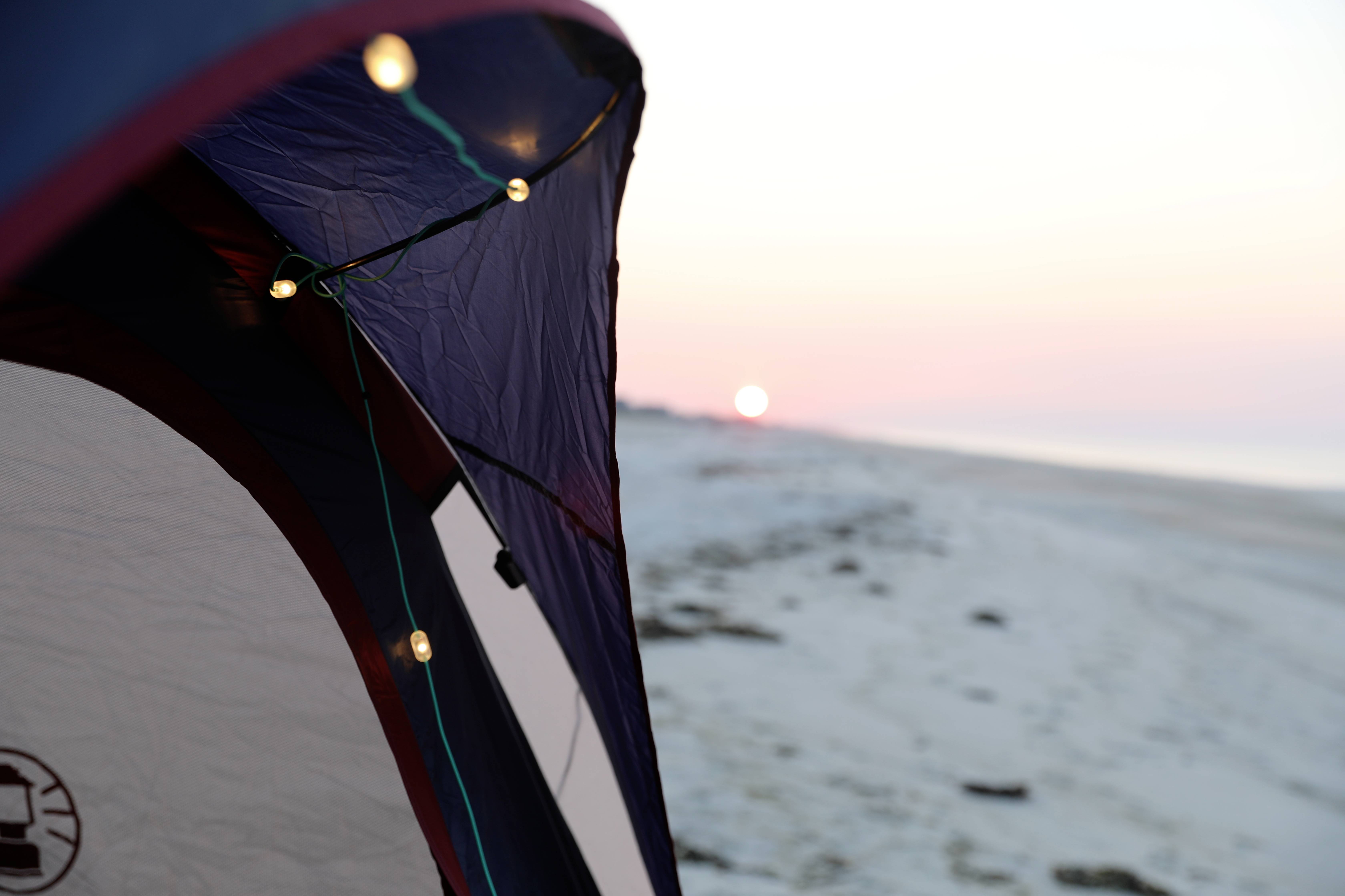 Tent met lichtsnoer op het strand bij zonsondergang; warme kleuren op de achtergrond, vredige en ontspannen sfeer.
