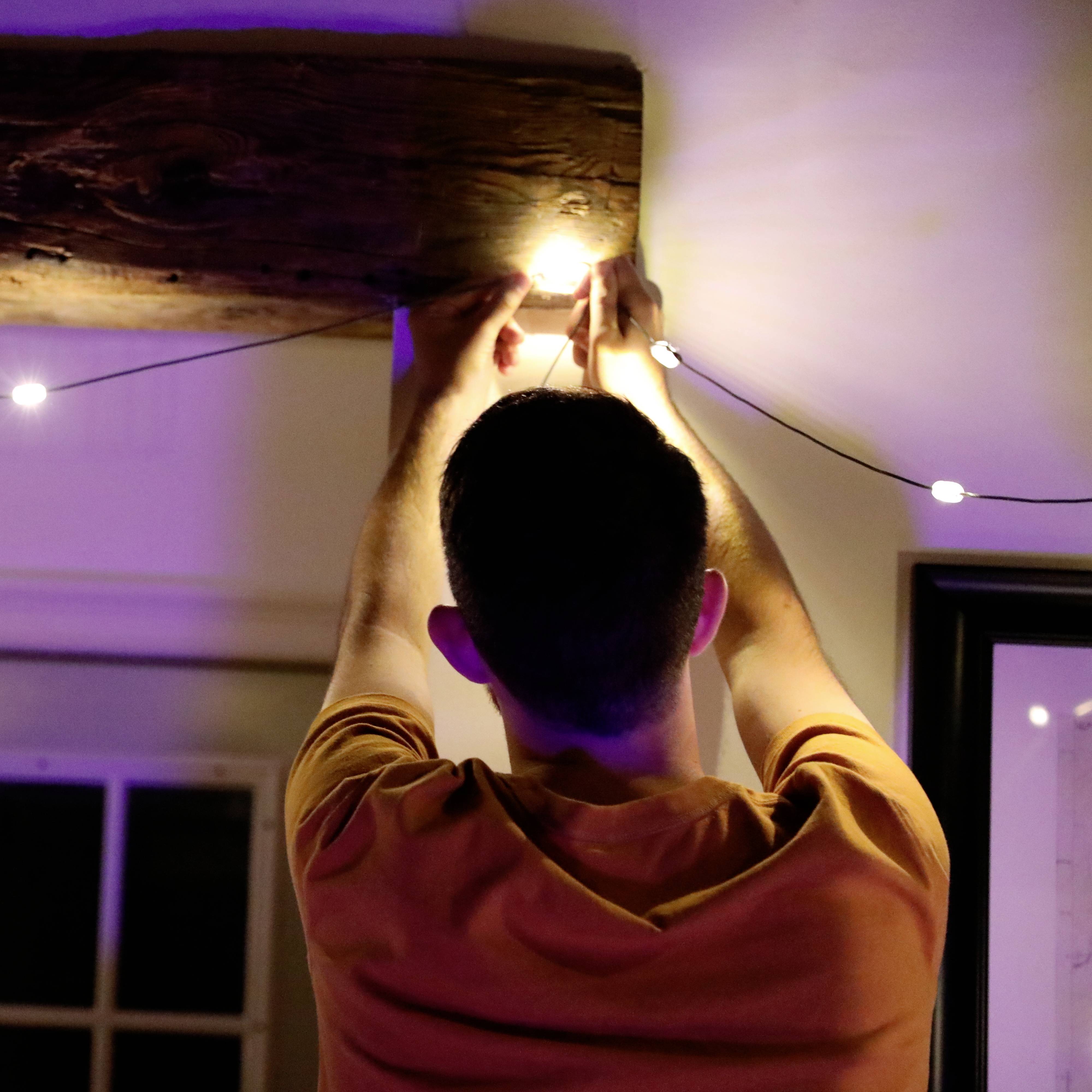 Een persoon repareert of monteert een gloeilamp aan het plafond. Op de achtergrond is een houten balk zichtbaar.